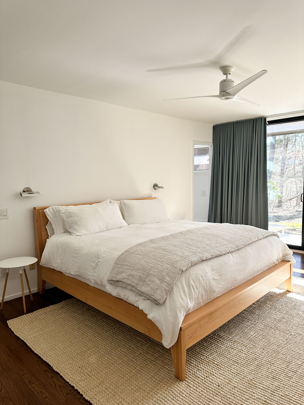 Spacious bedroom with a light wood platform bed, white bedding, and a light gray throw blanket. It features a ceiling fan, wall-mounted lights, and large sliding glass doors leading to an outdoor area.