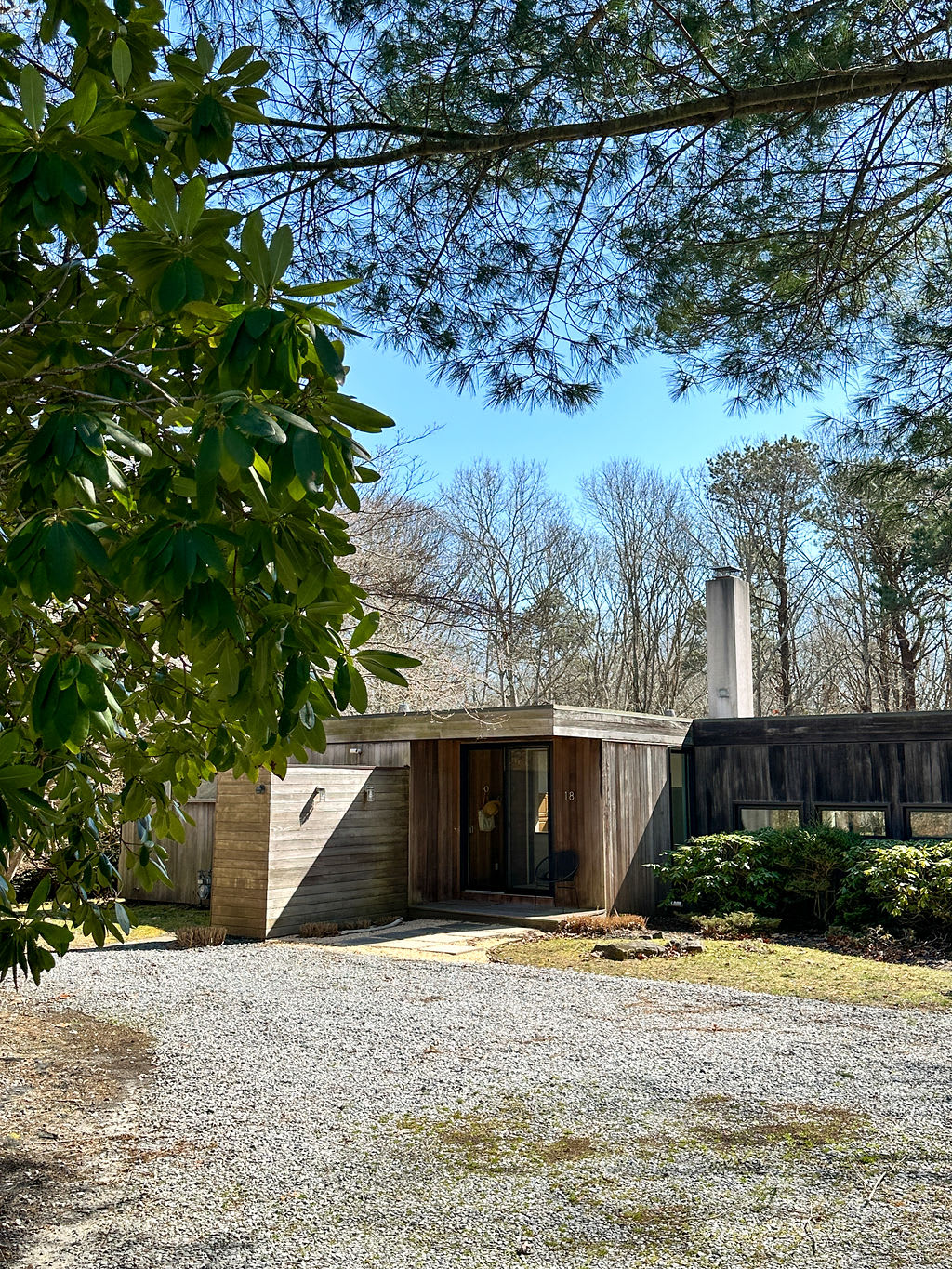 The exterior of a modern, wood-sided house with a gravel driveway, surrounded by trees and natural landscaping under a clear blue sky. The front entrance with its sliding glass door is visible.