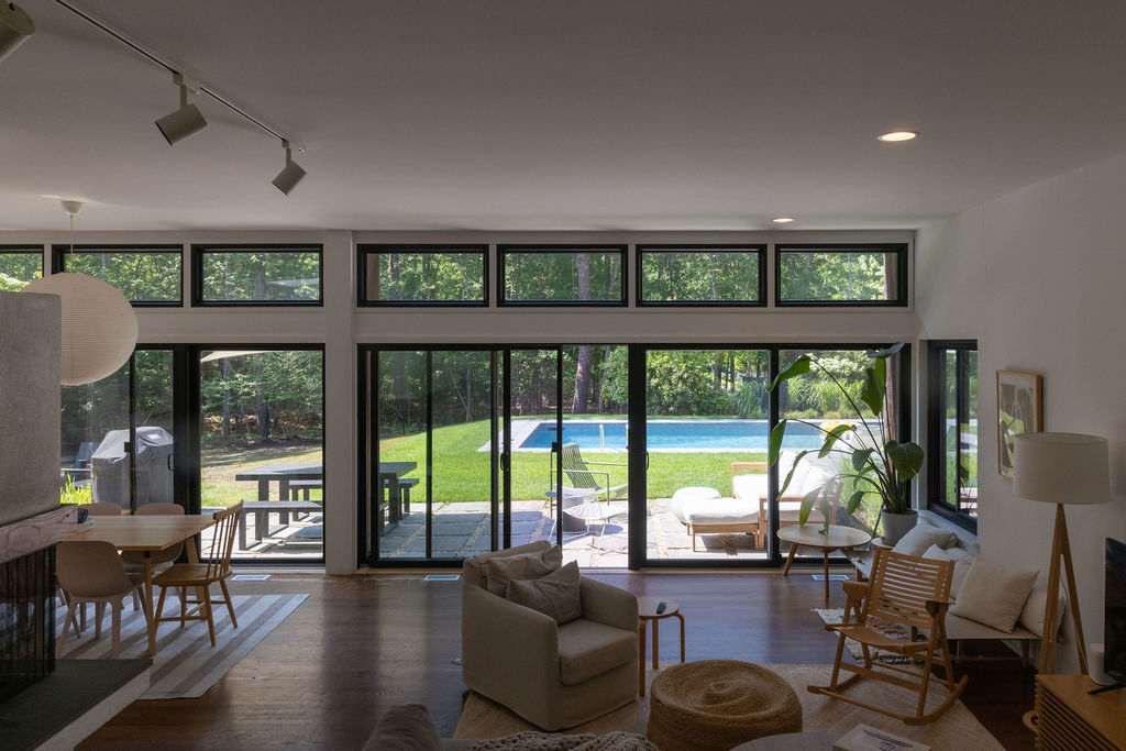 Open-concept living and dining area with hardwood floors, featuring a view of the outdoor pool through a wall of black-framed sliding glass doors and transom windows. The space is furnished with a sectional sofa and a dining table.