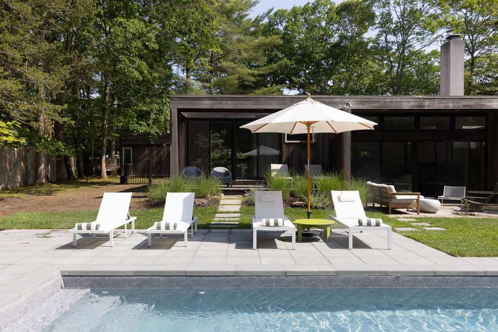 Modern outdoor pool area with white lounge chairs and a yellow side table, set on a light gray patio. The contemporary house exterior is visible in the background, surrounded by lush greenery.