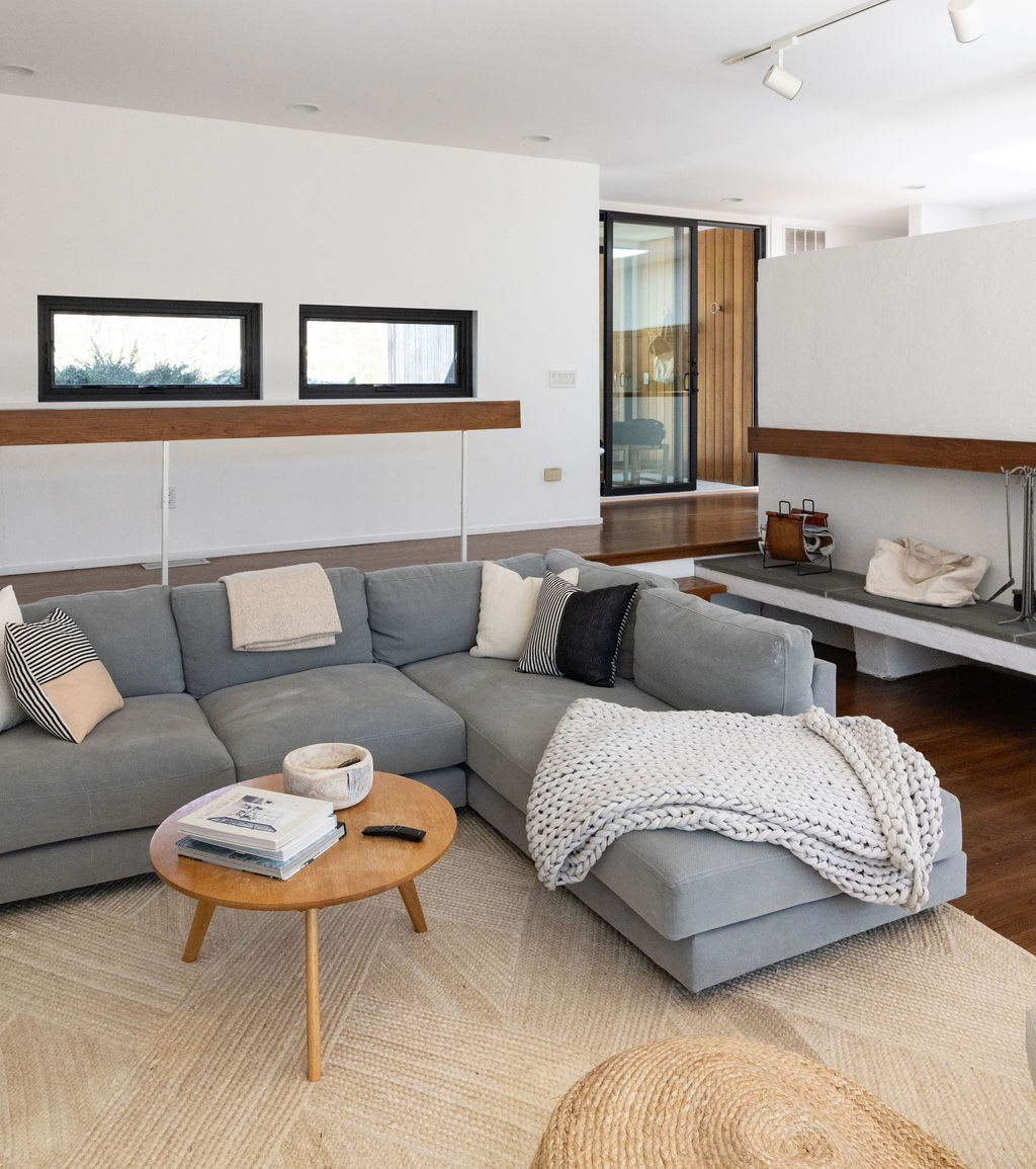 A detailed view of the living room's grey sectional sofa, adorned with various throw pillows and a chunky knit blanket, positioned in front of a modern fireplace with a concrete hearth and a wooden mantel.