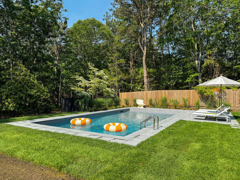 Outdoor swimming pool with a light gray paved surround, flanked by a manicured lawn and tall trees. White lounge chairs and an umbrella are positioned poolside, with floating rings in the water.
