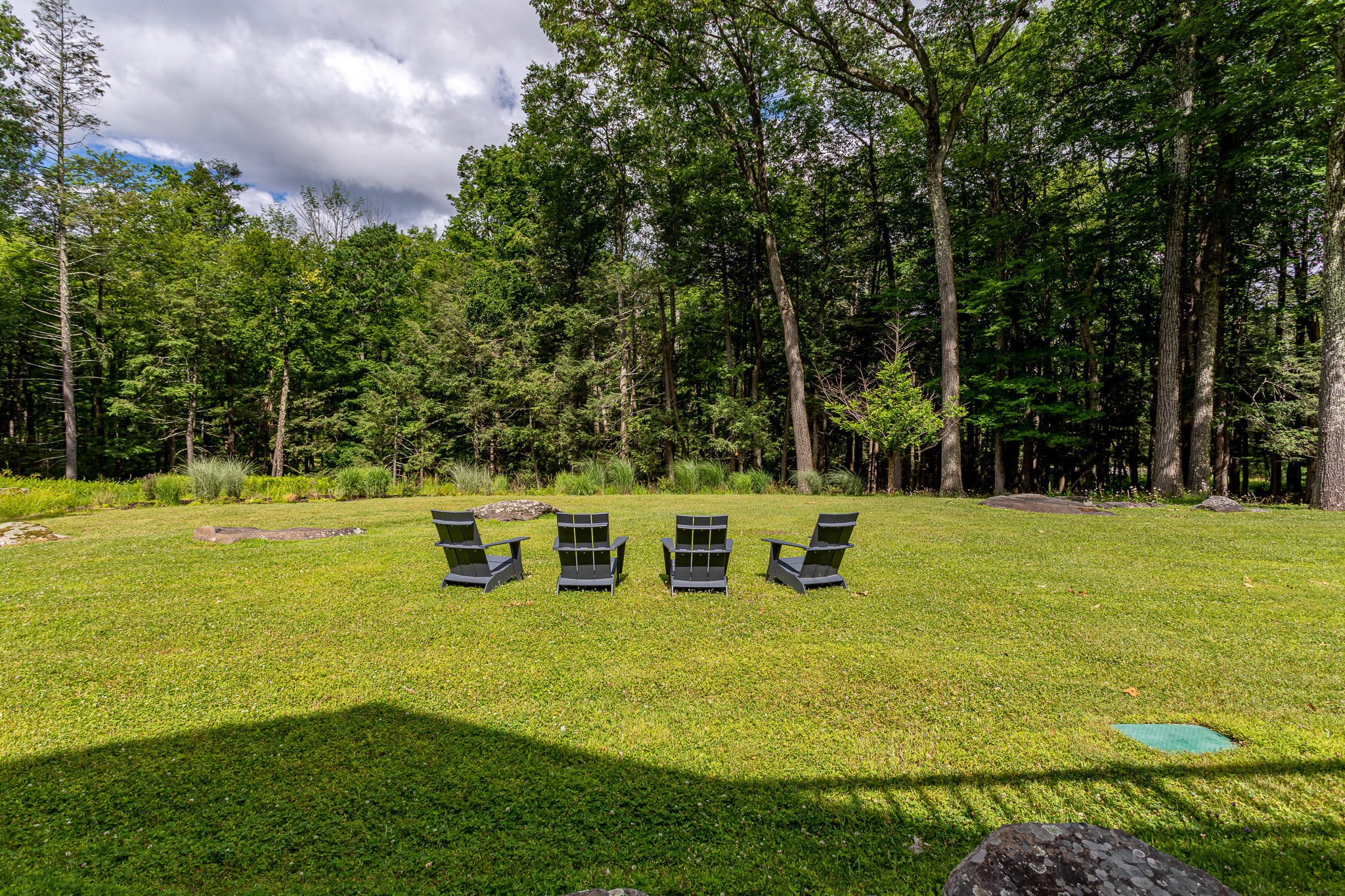 Expansive green lawn with four dark Adirondack chairs arranged in a semicircle, surrounded by lush green trees under a partly cloudy sky, offering a serene outdoor seating area.