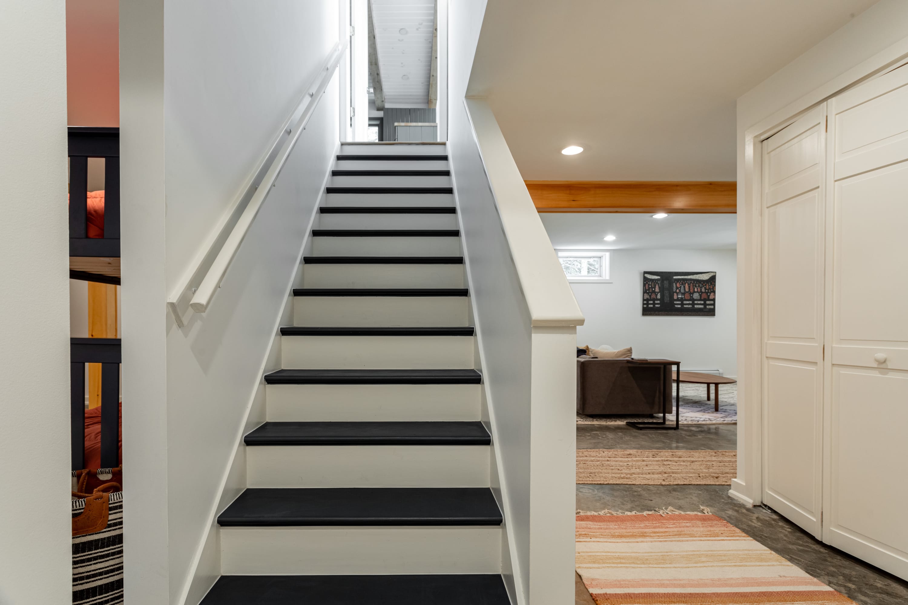 Bright white staircase with contrasting black treads and a white railing, leading upwards. A glimpse of a bunk room is visible to the left, and a living area with a rug and wooden beam is to the right.