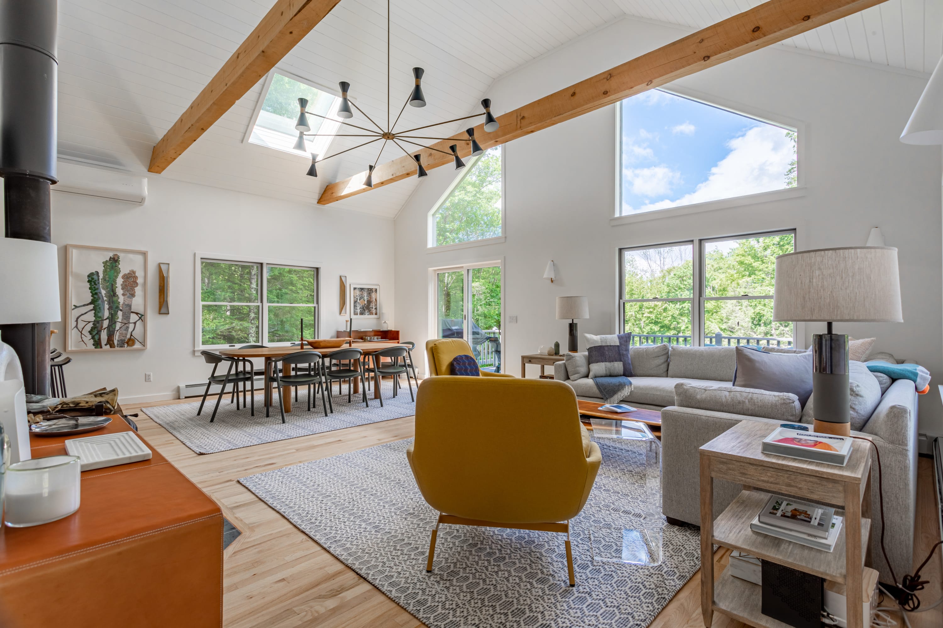 A bright, open-concept living and dining area features a vaulted white ceiling with exposed wooden beams, large windows offering lush green views, and a modern chandelier over a wooden dining table.