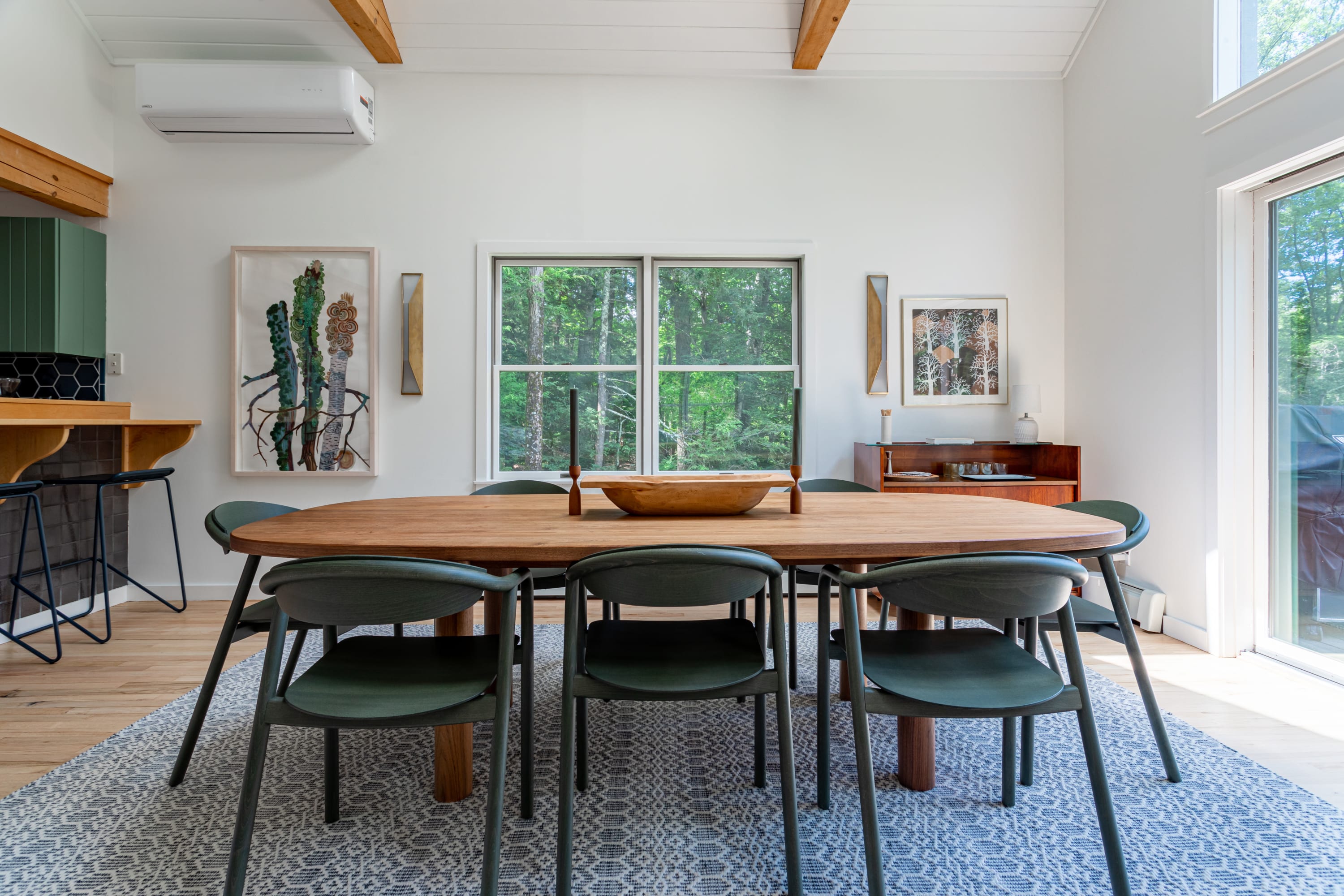 Elegant dining area featuring a large oval wooden table with dark green chairs, set on a patterned rug, with a window overlooking the forest and a view into the adjacent kitchen.