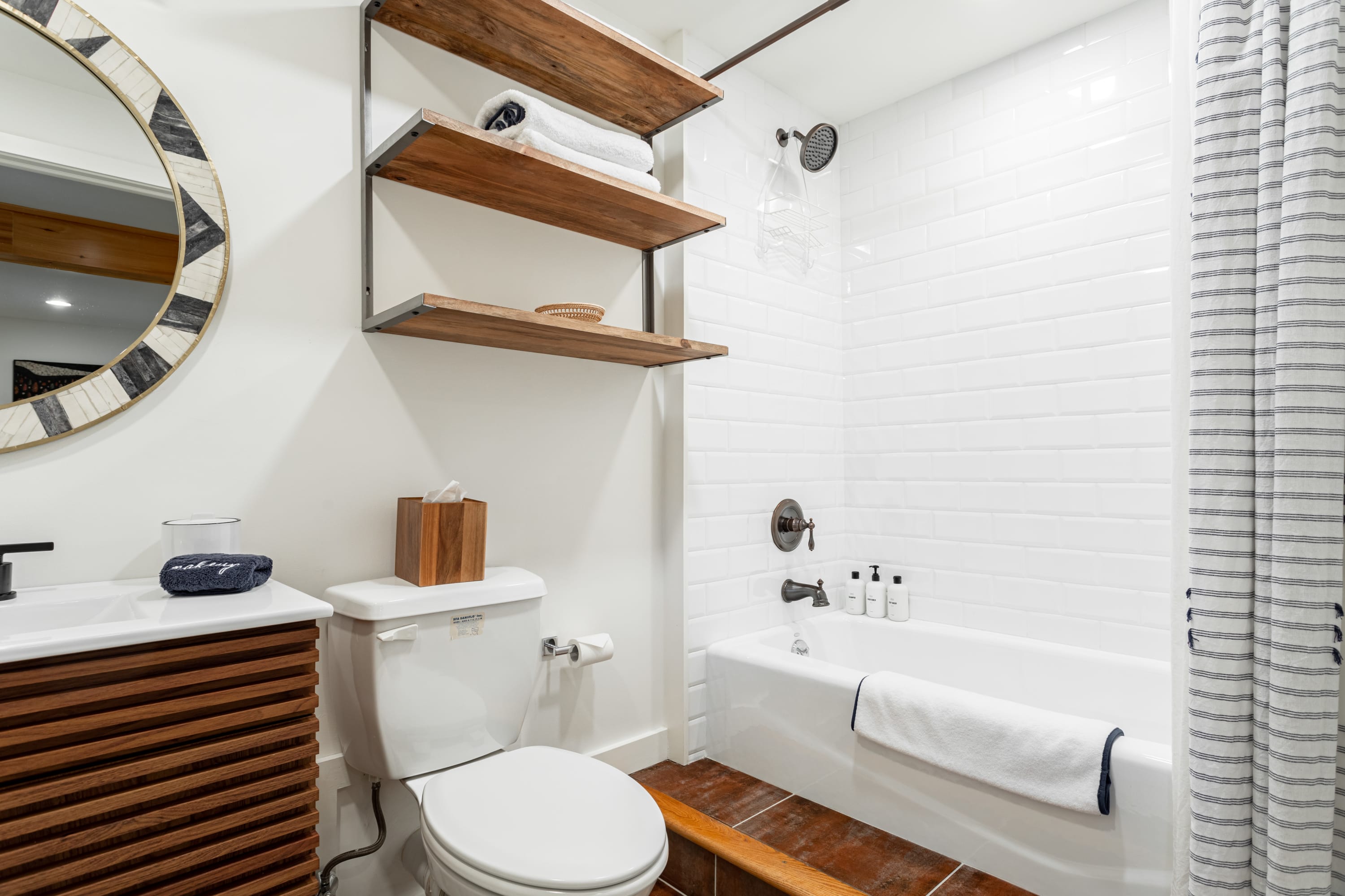 Bathroom with a white subway-tiled shower/tub combination, a toilet, and a vanity featuring a slatted wooden cabinet and a round mirror. Rustic wooden shelves with towels are mounted above the toilet.
