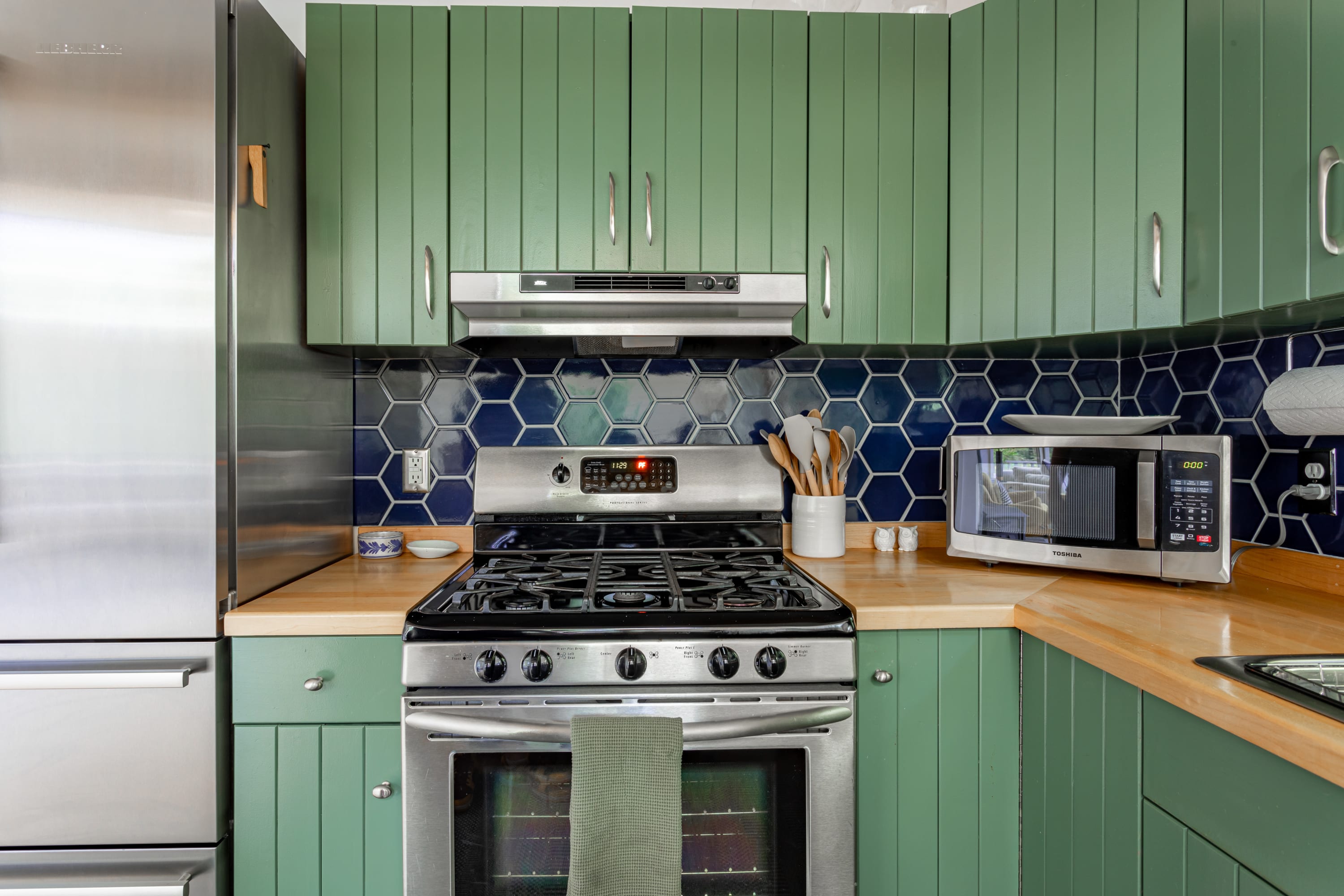A close-up of the kitchen shows a stainless steel gas range with an overhead microwave, set against a striking dark blue hexagonal tile backsplash and light wood countertops, flanked by green cabinets.