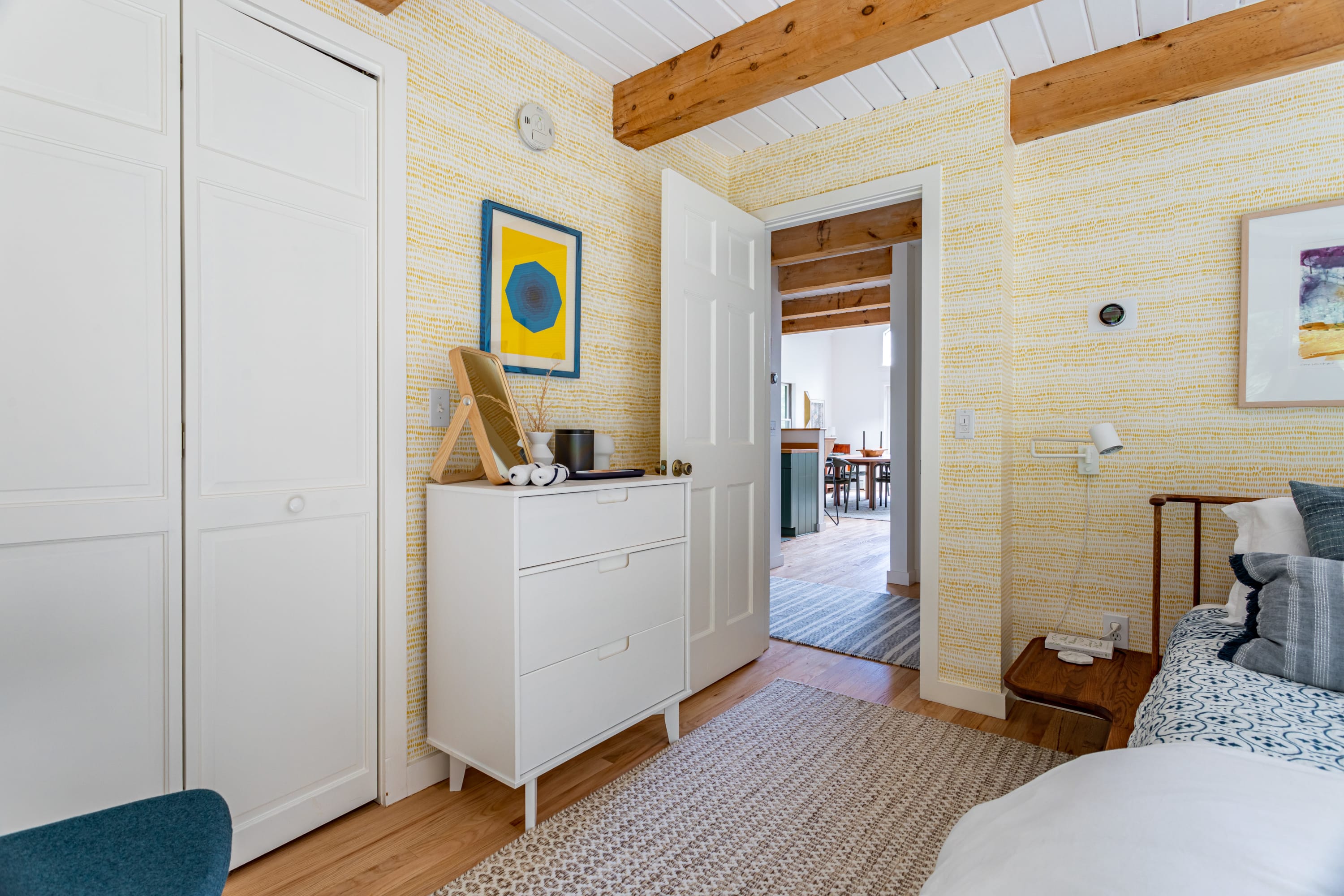 View of bedroom-1 showing a white dresser with a standing mirror, white bi-fold closet doors, and the distinctive yellow patterned wallpaper. An open doorway leads to an adjacent room.