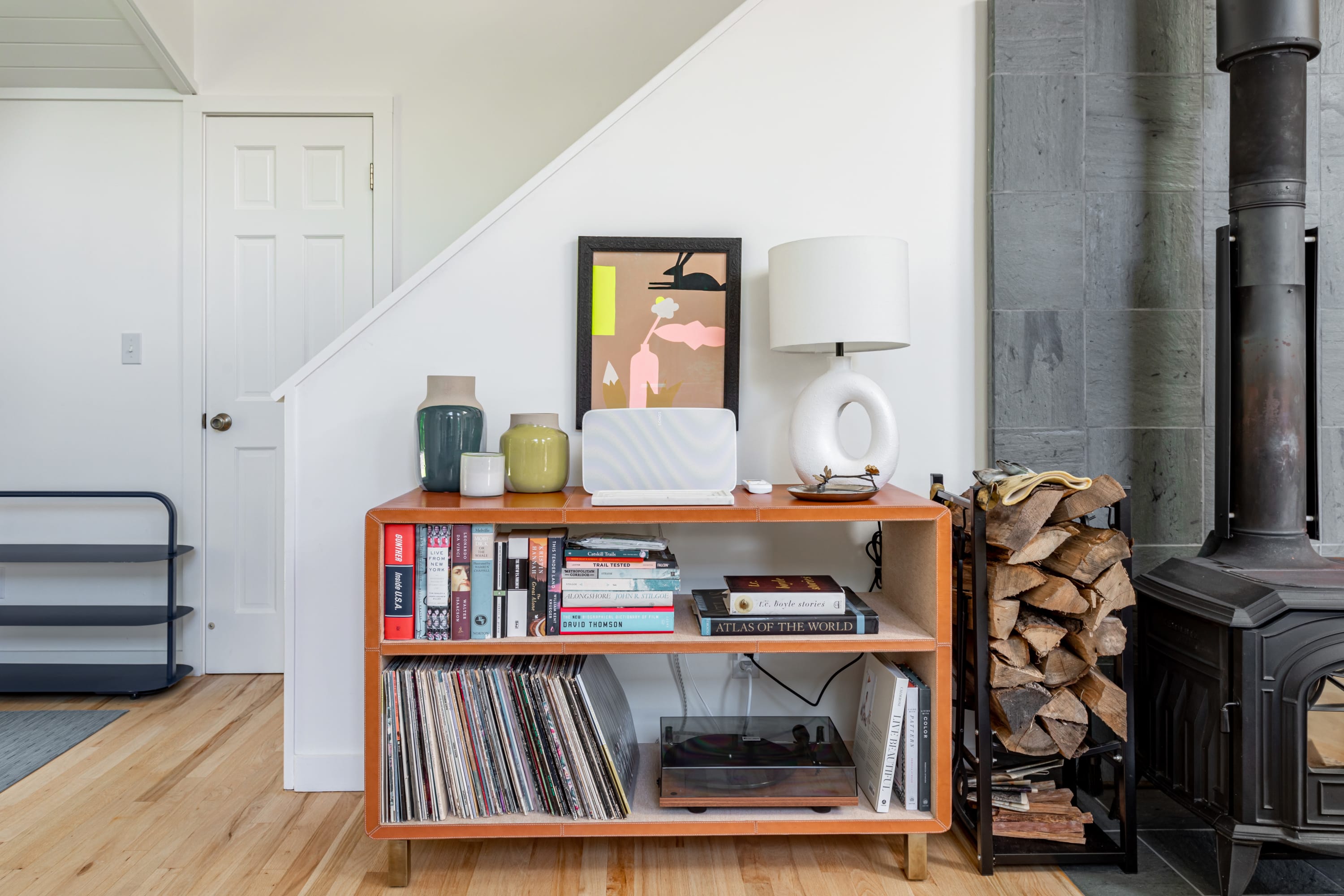 A stylish entryway features a wooden console table adorned with books, records, and decorative items, next to a black wood-burning stove with stacked firewood, leading to a white staircase.