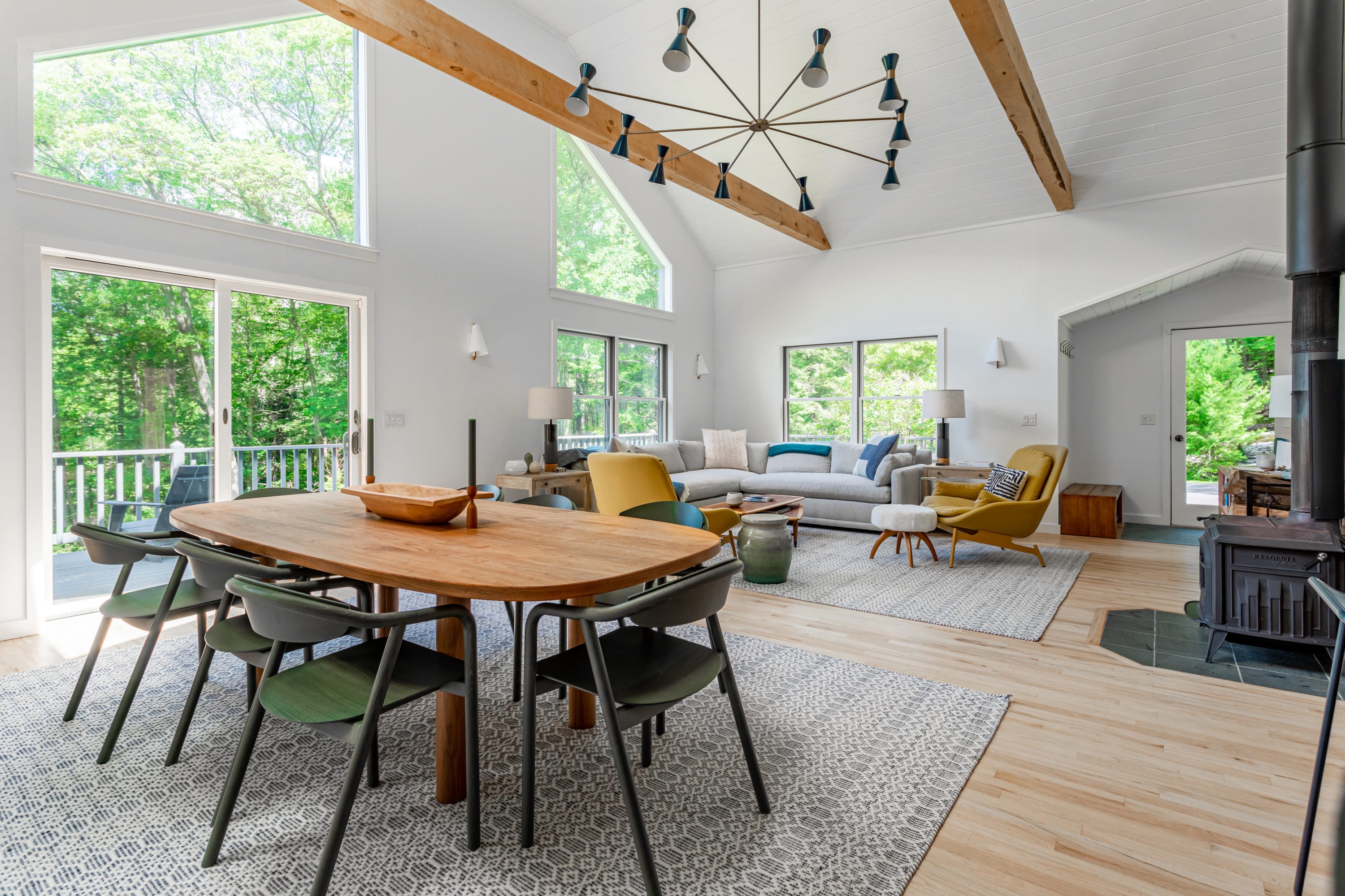 An expansive open-plan living and dining space boasts a vaulted ceiling with exposed wooden beams, a striking modern chandelier, and large windows and sliding doors that open to a deck, offering abundant natural light and forest views.