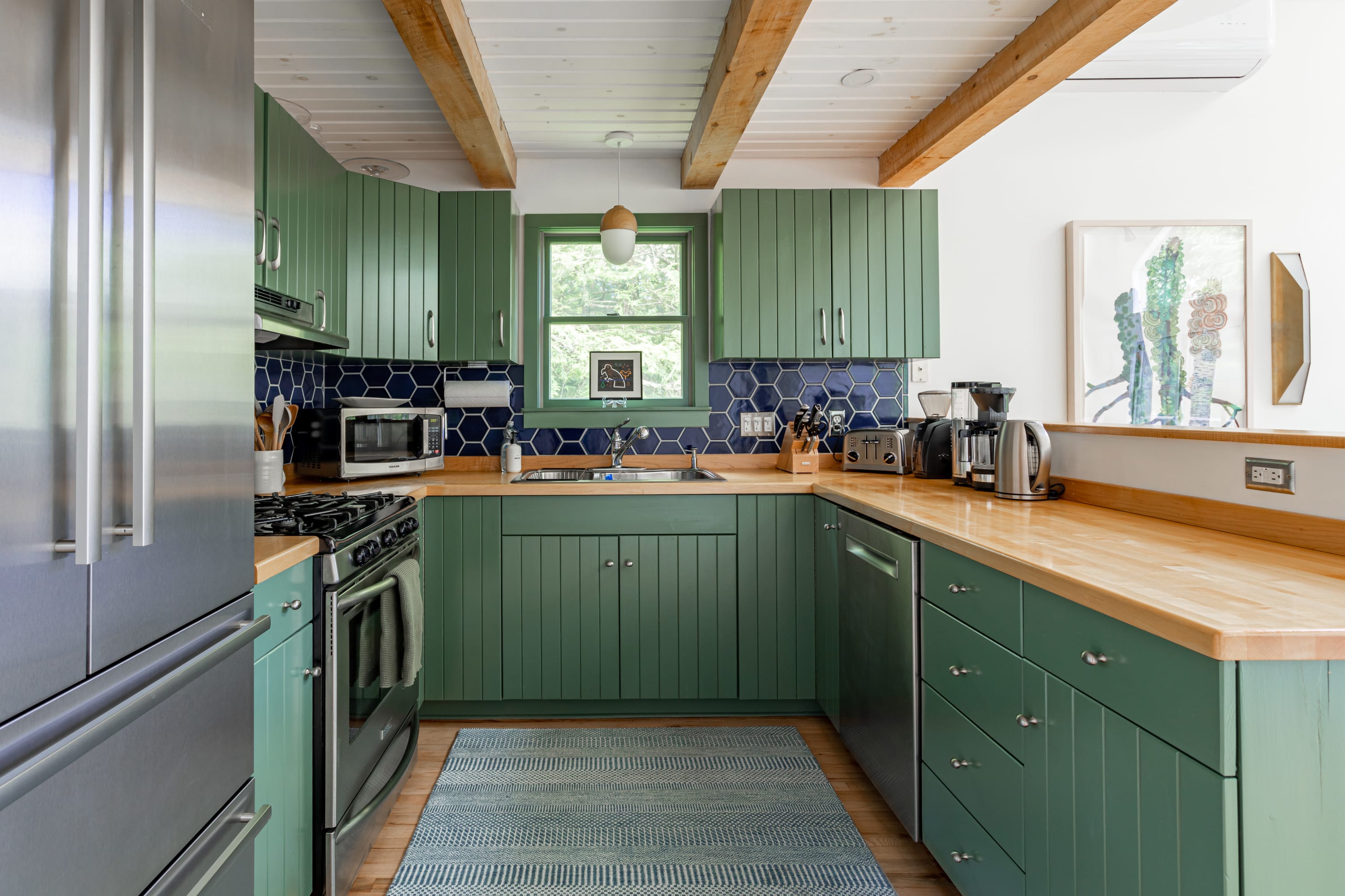 A compact yet functional kitchen features vibrant green cabinetry, light wood countertops, and a dark blue hexagonal tile backsplash, equipped with stainless steel appliances and a window overlooking the lush outdoors.