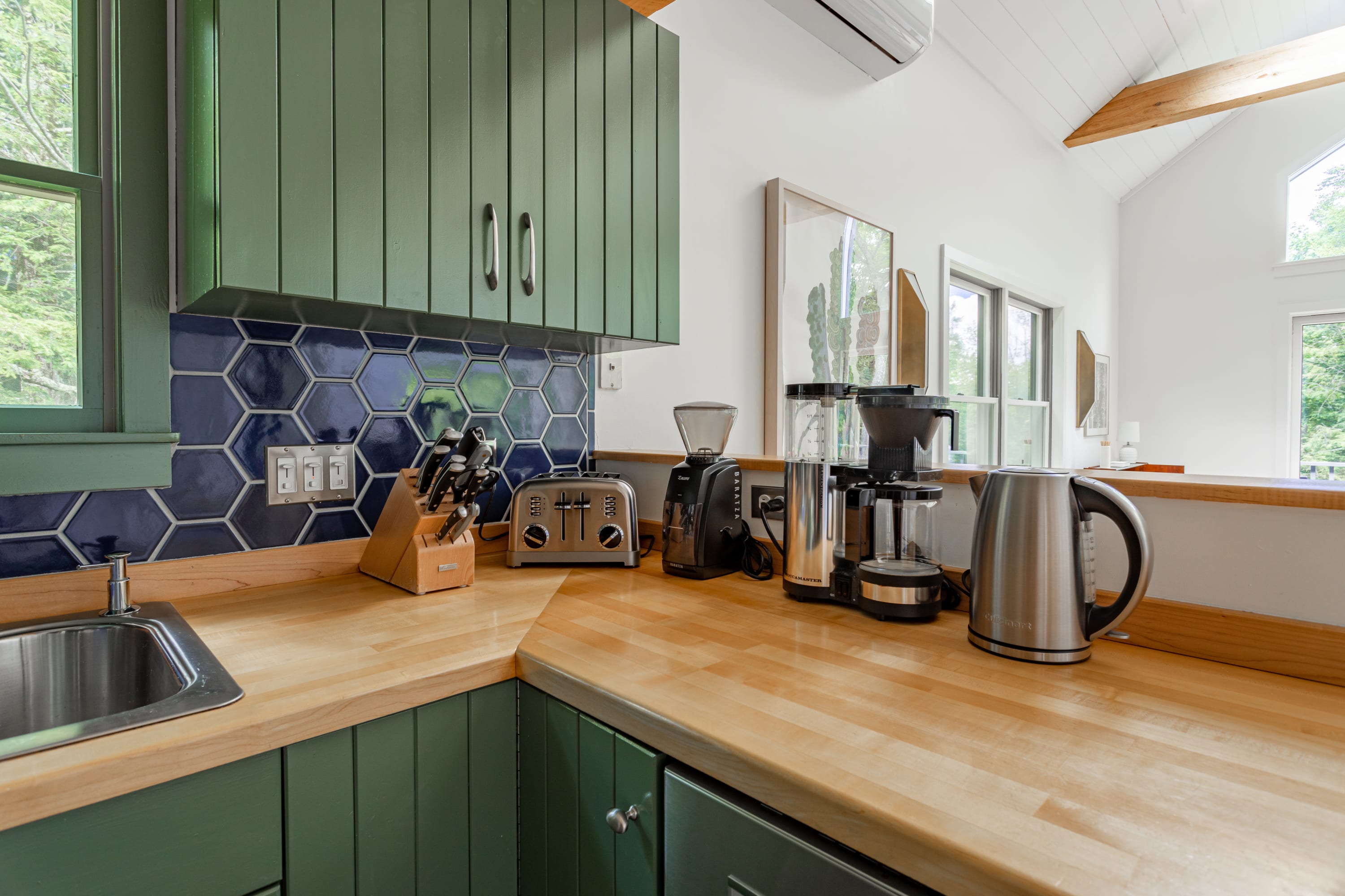 A section of the kitchen countertop displays various coffee-making appliances, a toaster, and a knife block, all on light wood surfaces with a dark blue hexagonal tile backsplash and green cabinets.