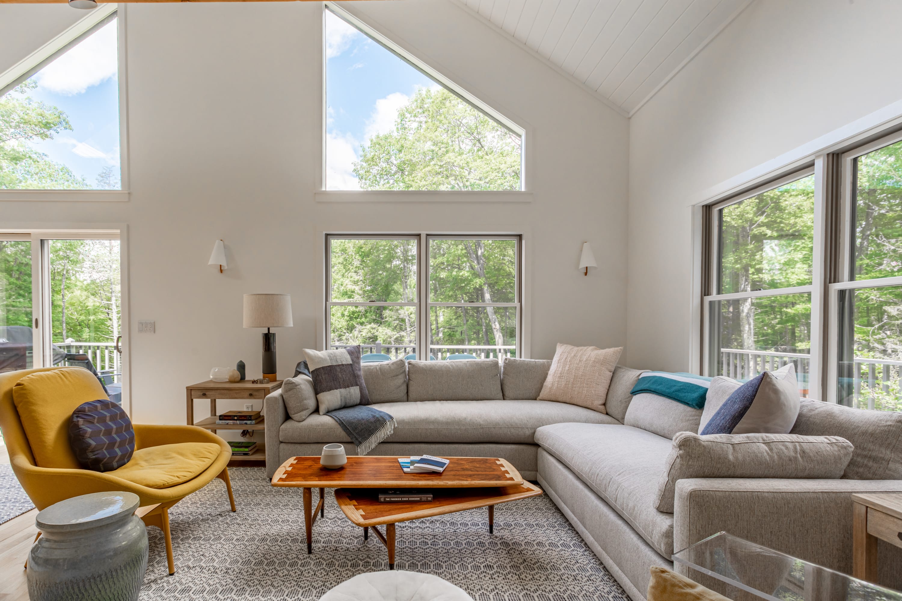 A cozy living room corner features a large L-shaped sectional sofa with throw pillows, a unique wooden coffee table, and a bright yellow armchair, all illuminated by natural light from expansive windows overlooking a green landscape.