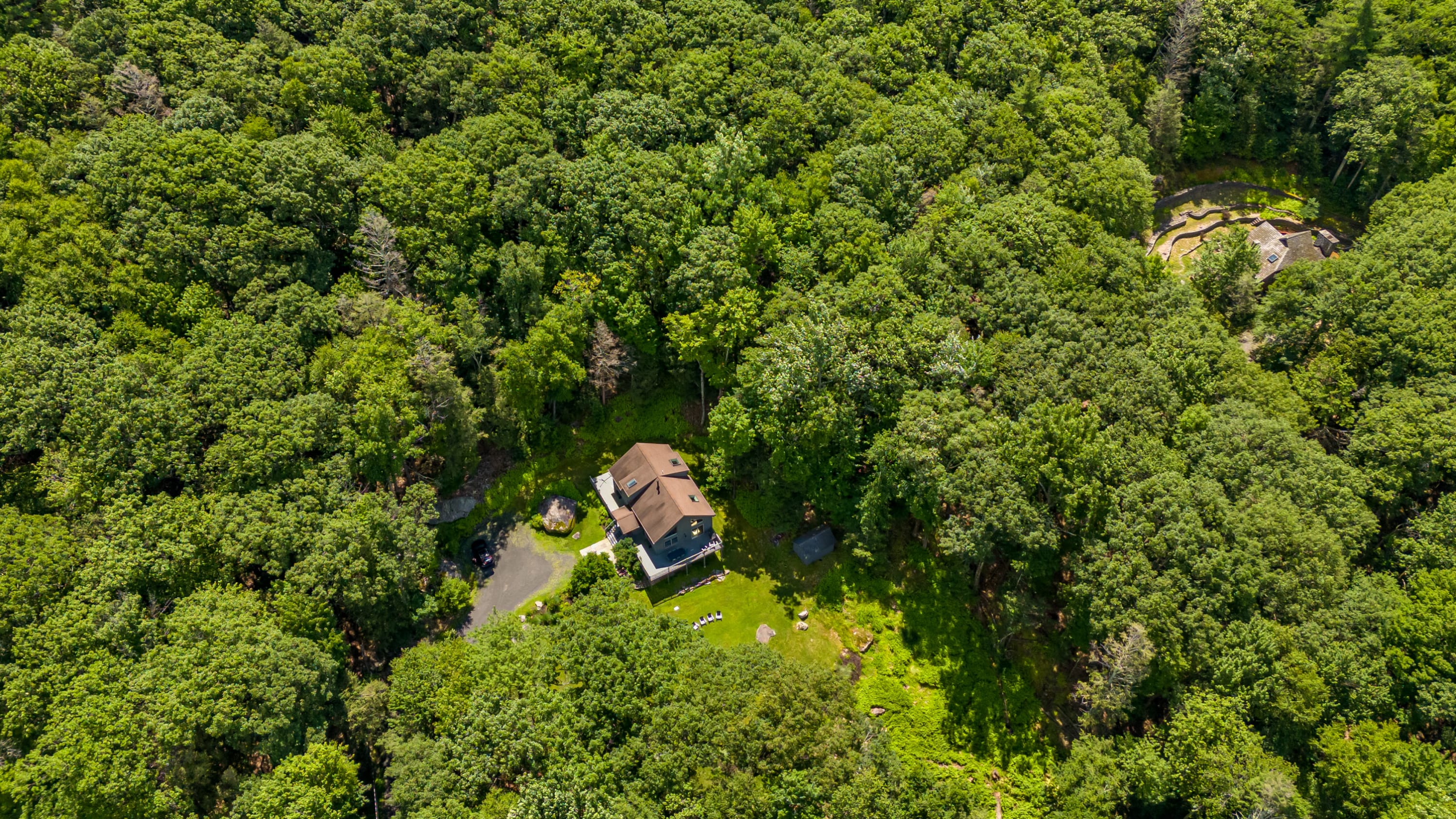 An aerial view showcases a modern chalet-style home with a brown roof nestled deep within a dense, vibrant green forest, highlighting its secluded and natural setting.