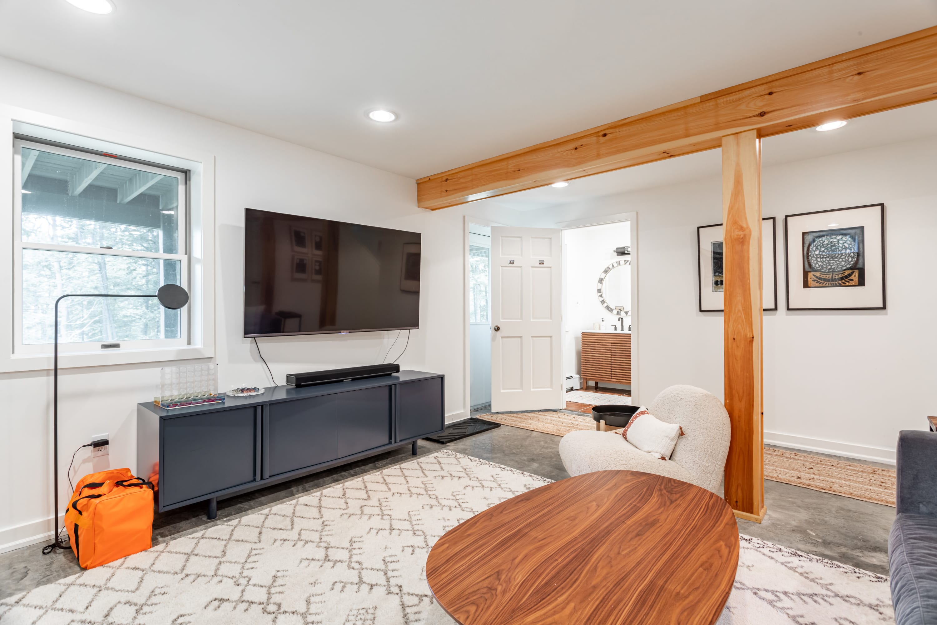Another view of the living room, showcasing a dark gray media console with a TV, a wooden coffee table, and a cream-colored armchair. A doorway leads to a bathroom, and a large wooden beam is a central architectural feature.