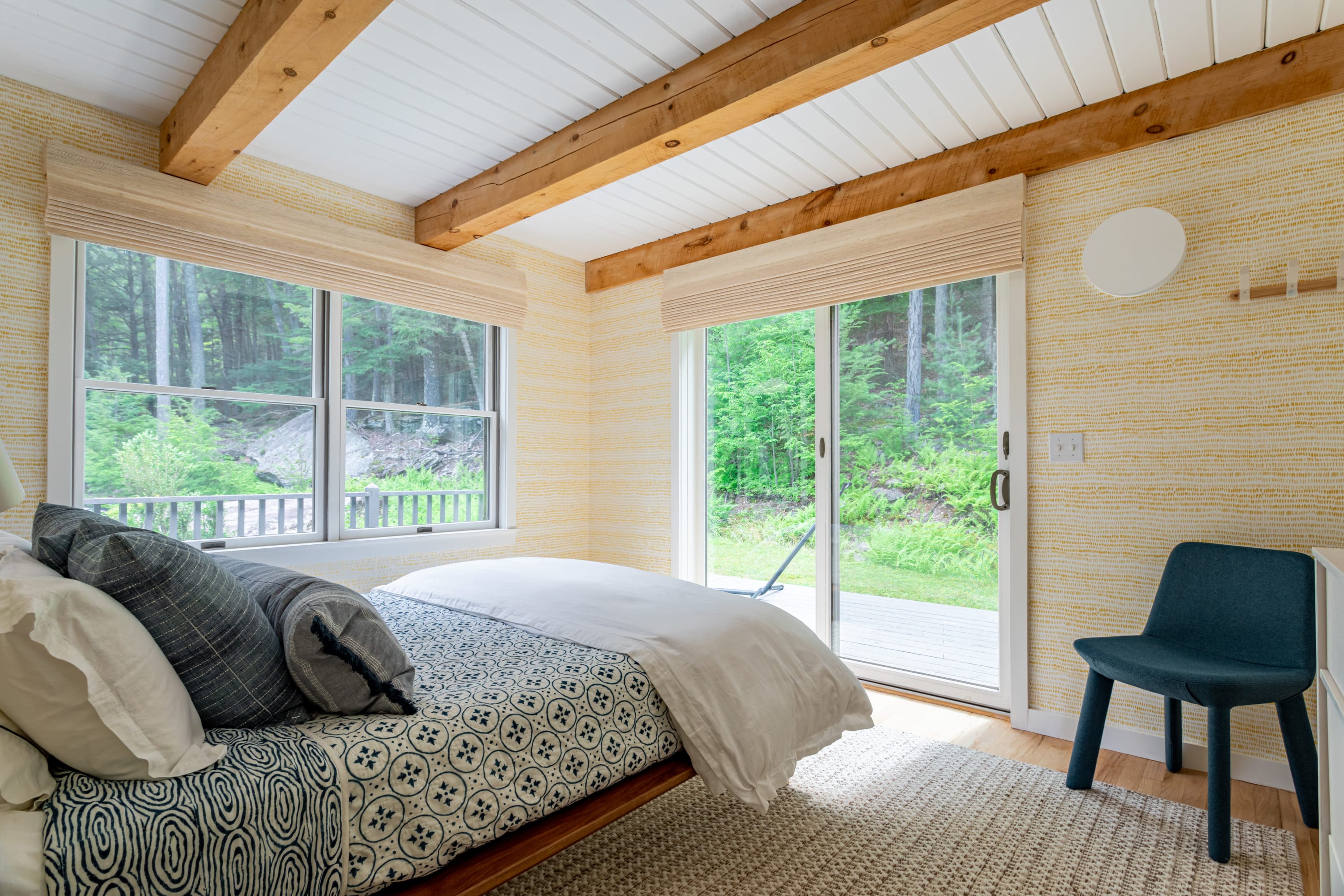 A bright bedroom features a bed with blue and white patterned bedding, yellow textured wallpaper, exposed wooden beams on a white ceiling, and large windows and a sliding door offering direct access to the outdoors and forest views.