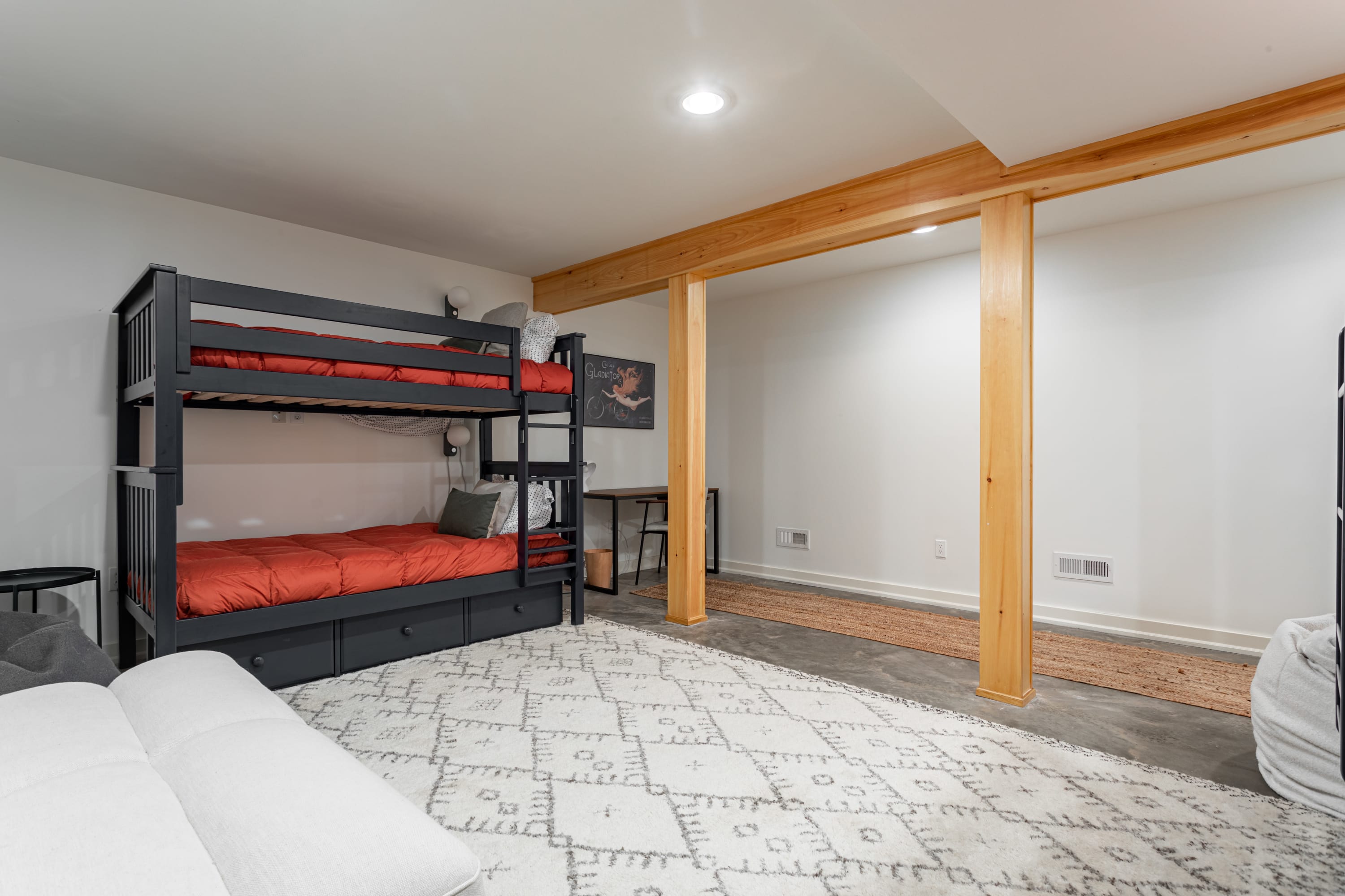 Another angle of the bunk room, highlighting a black bunk bed with orange bedding, a white sofa, and a dedicated desk area. Prominent wooden support beams and a long jute rug complement the concrete floor.