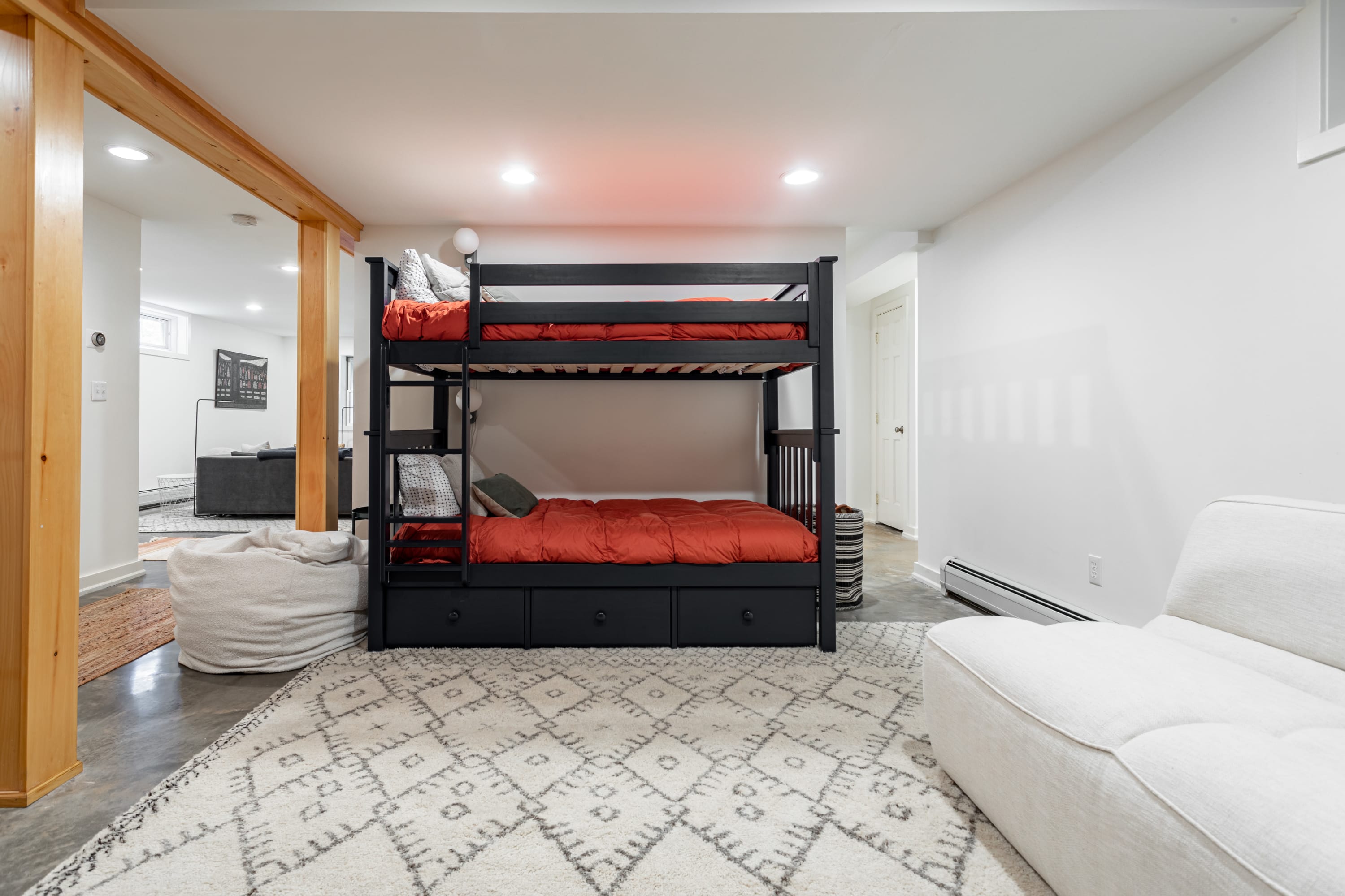 Close-up of a black bunk bed with vibrant orange bedding and integrated storage drawers, positioned on a patterned rug. A white sofa is partially visible, and wooden beams frame the sleeping area.