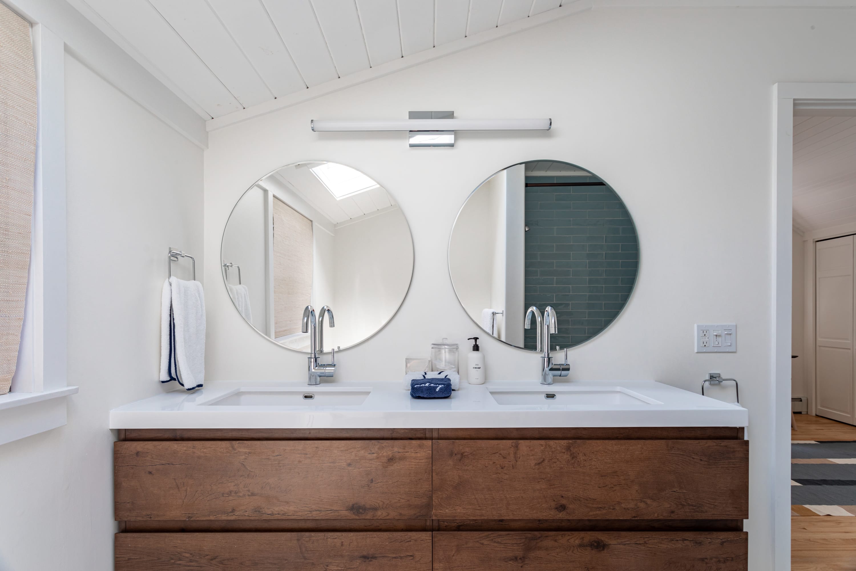 Modern bathroom featuring a double vanity with a rich wooden cabinet, white countertop, two round mirrors, and a sleek linear light fixture. The space has white walls and a vaulted, planked ceiling.
