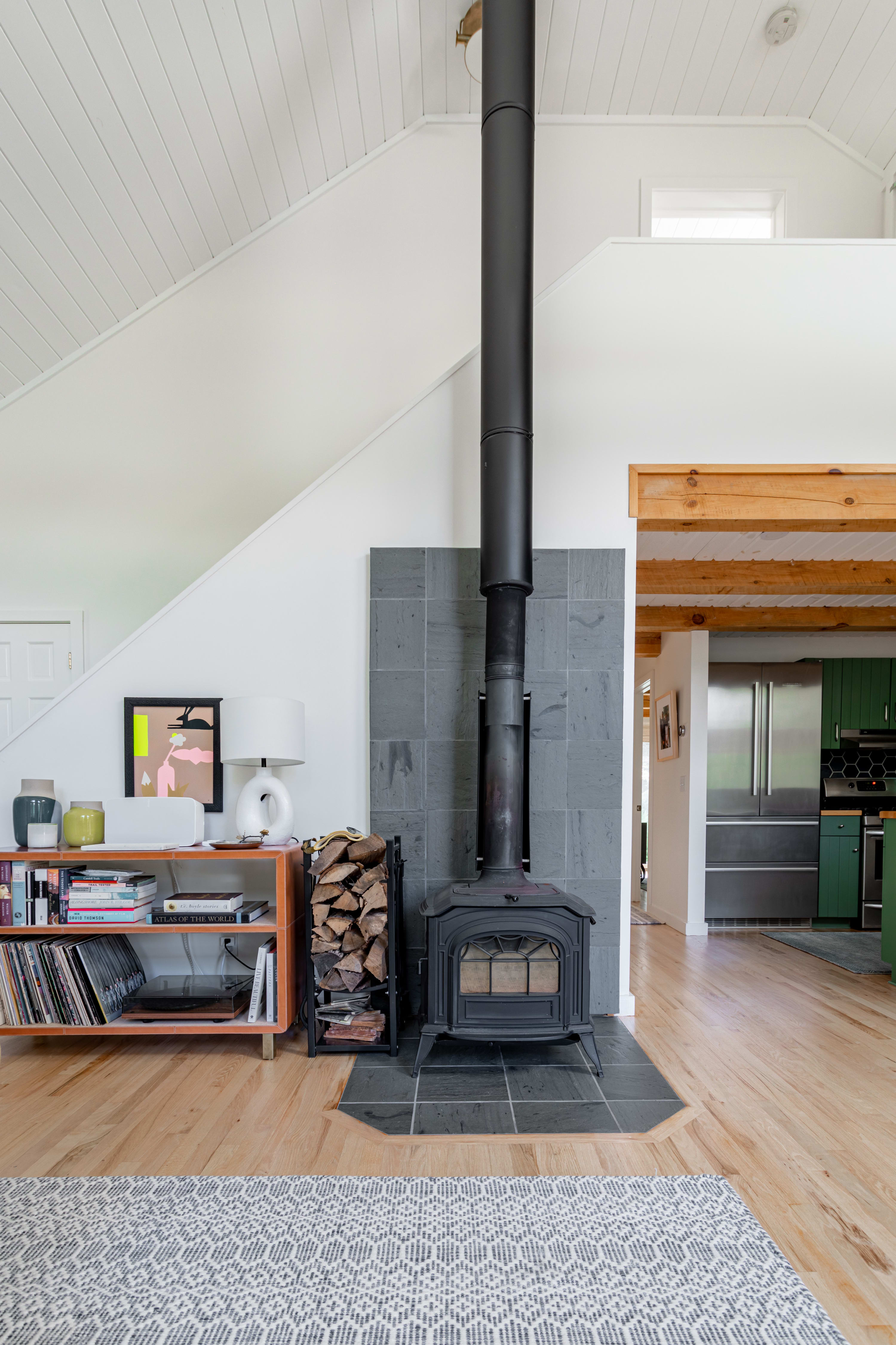 Detail shot of the living room showcasing a black wood-burning stove on a slate base, flanked by a modern orange bookshelf filled with records and decor, and stacked firewood.
