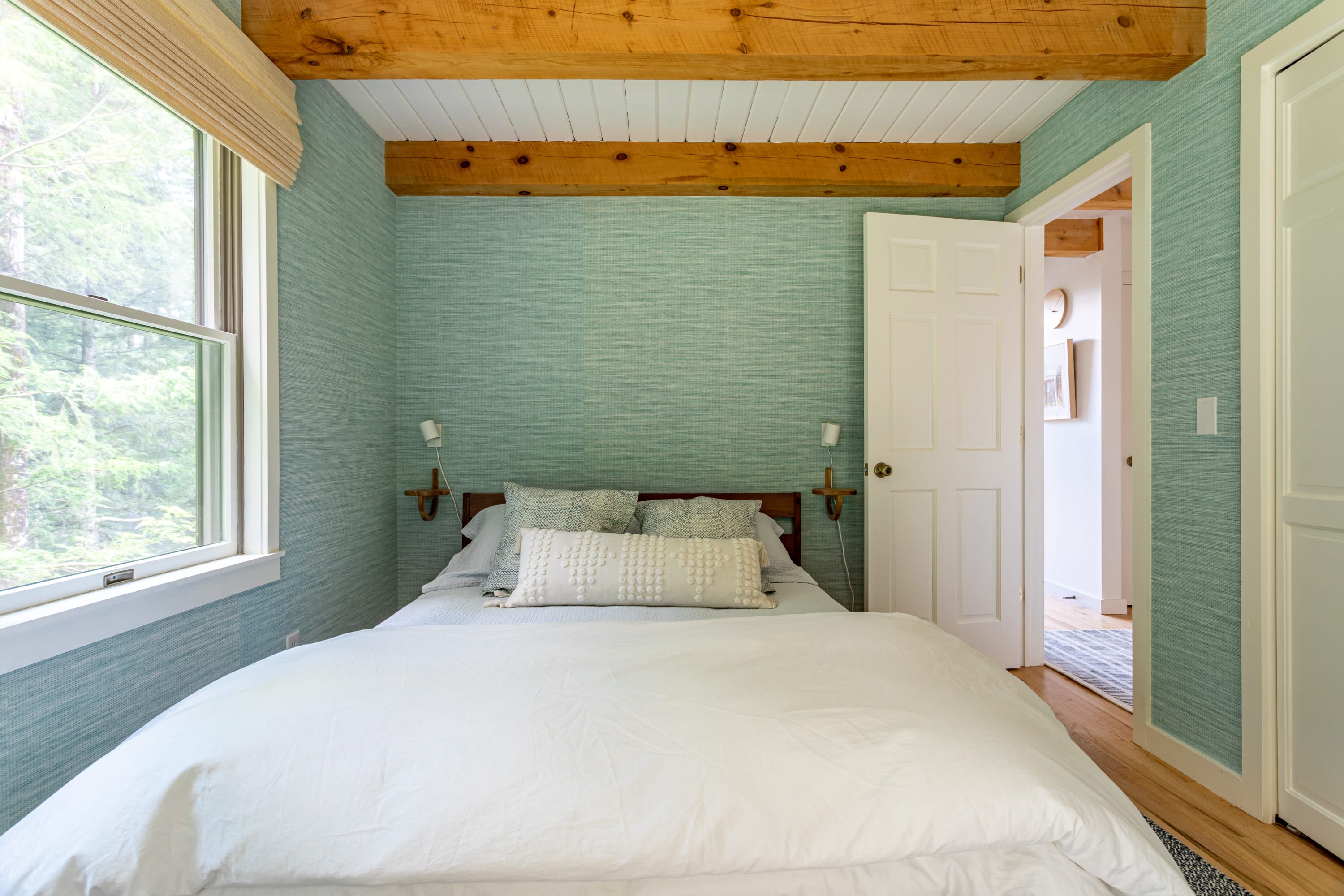 Head-on view of bedroom-2, featuring a bed with white bedding, light blue textured wallpaper, and exposed wooden ceiling beams. A window on the left overlooks trees, and a closet is visible on the right.