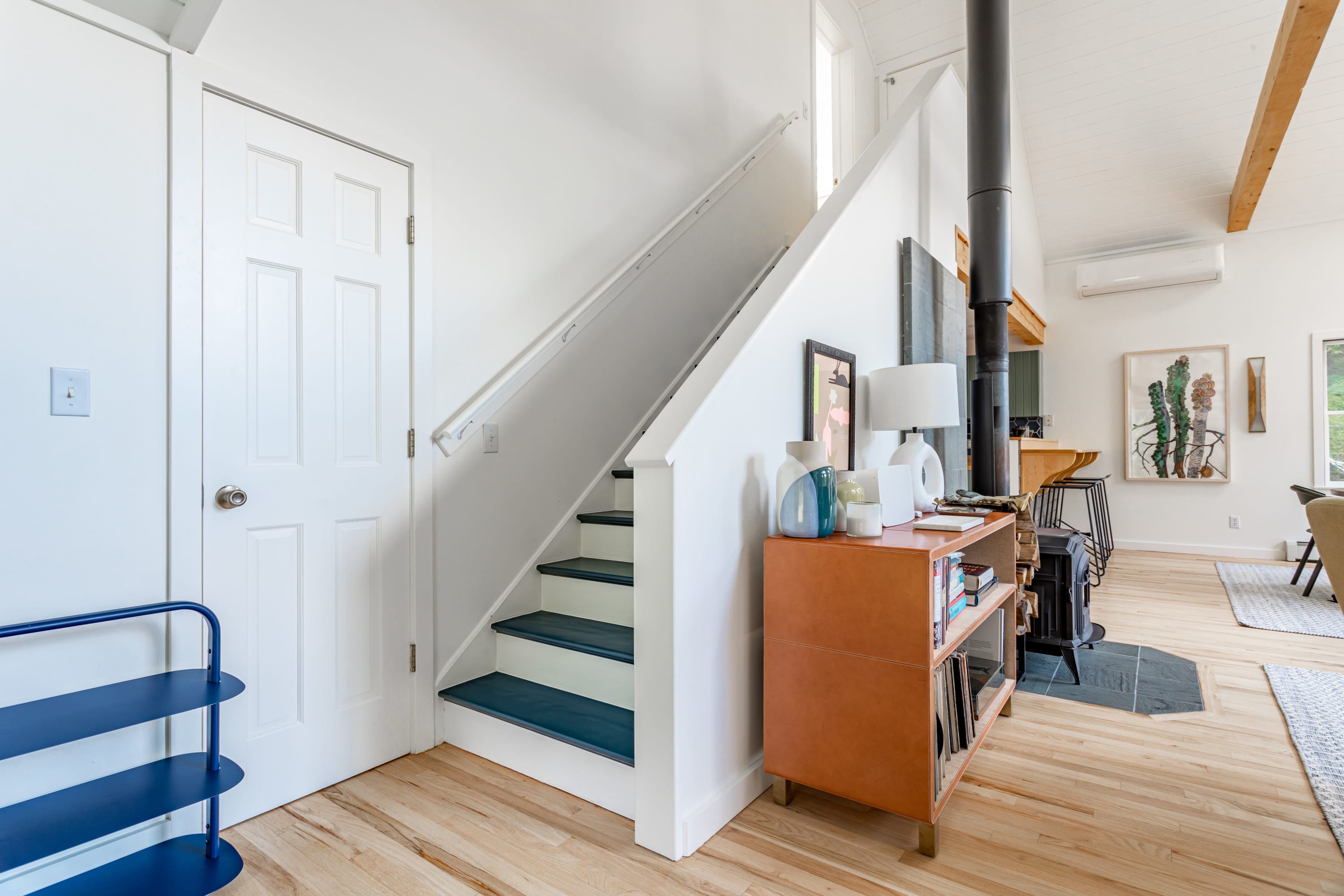 A bright foyer area with white walls and a staircase featuring dark blue risers. A wooden console table with decorative items is visible, leading into an open living space.