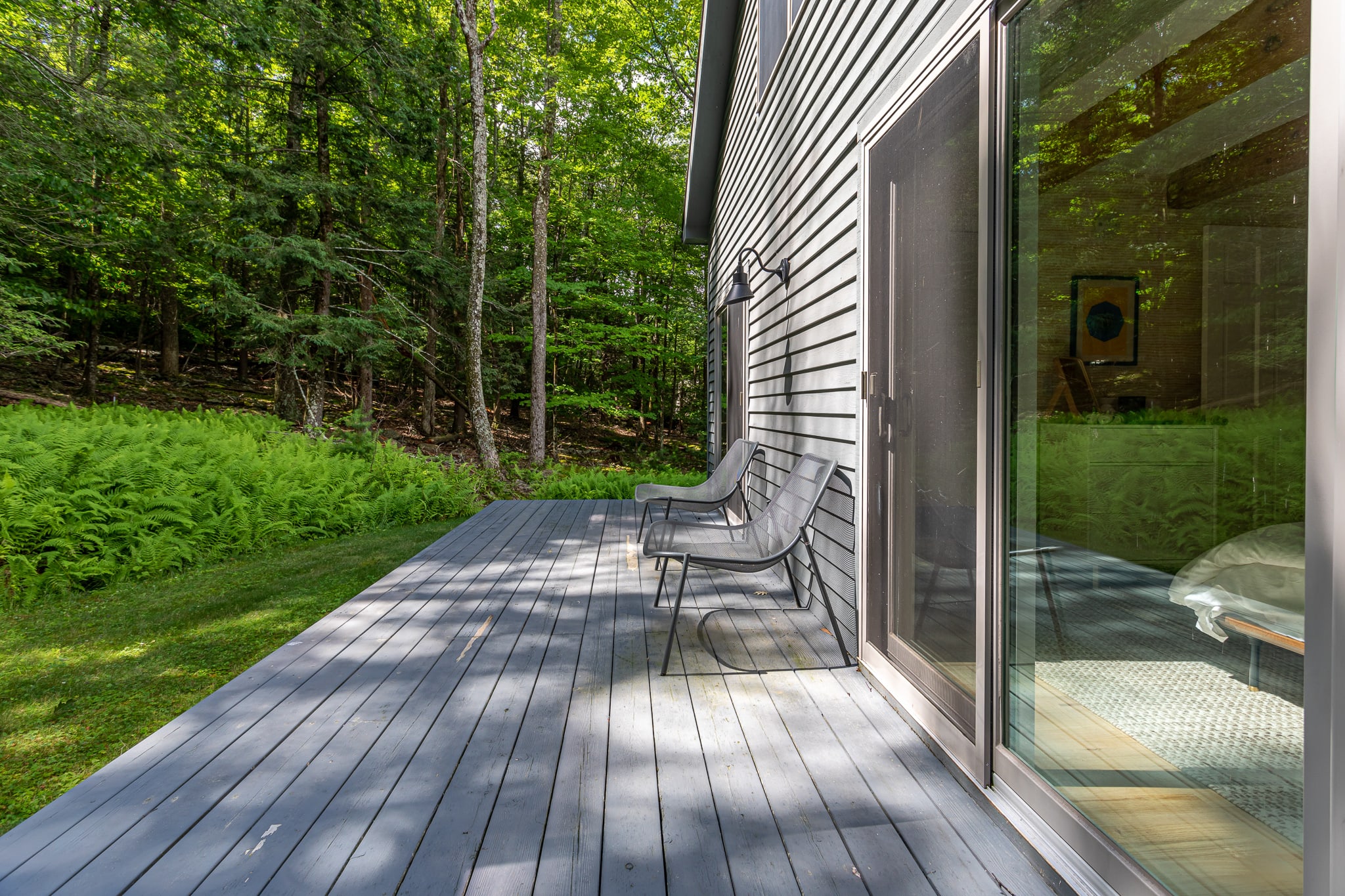 A serene section of the gray composite deck, featuring two modern dark gray lounge chairs positioned near a large sliding glass door. The deck is bordered by vibrant green ferns and dense forest, offering a private outdoor retreat.