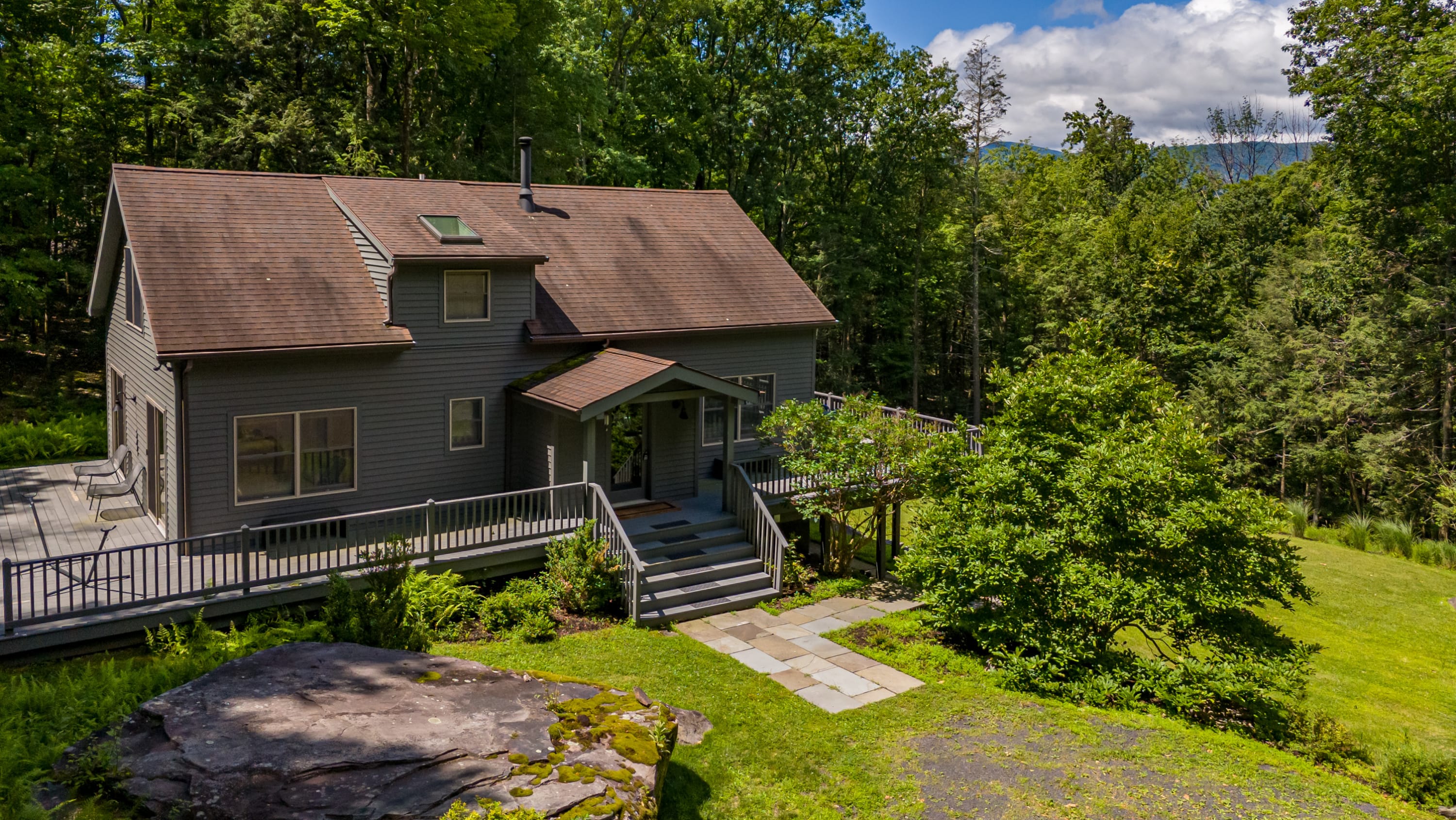 An elevated view of the gray chalet-style house, showcasing a multi-level deck with a covered entrance and stone pathway leading to the front door. The property is surrounded by lush green trees and a large rock formation, with a glimpse of distant mountains.