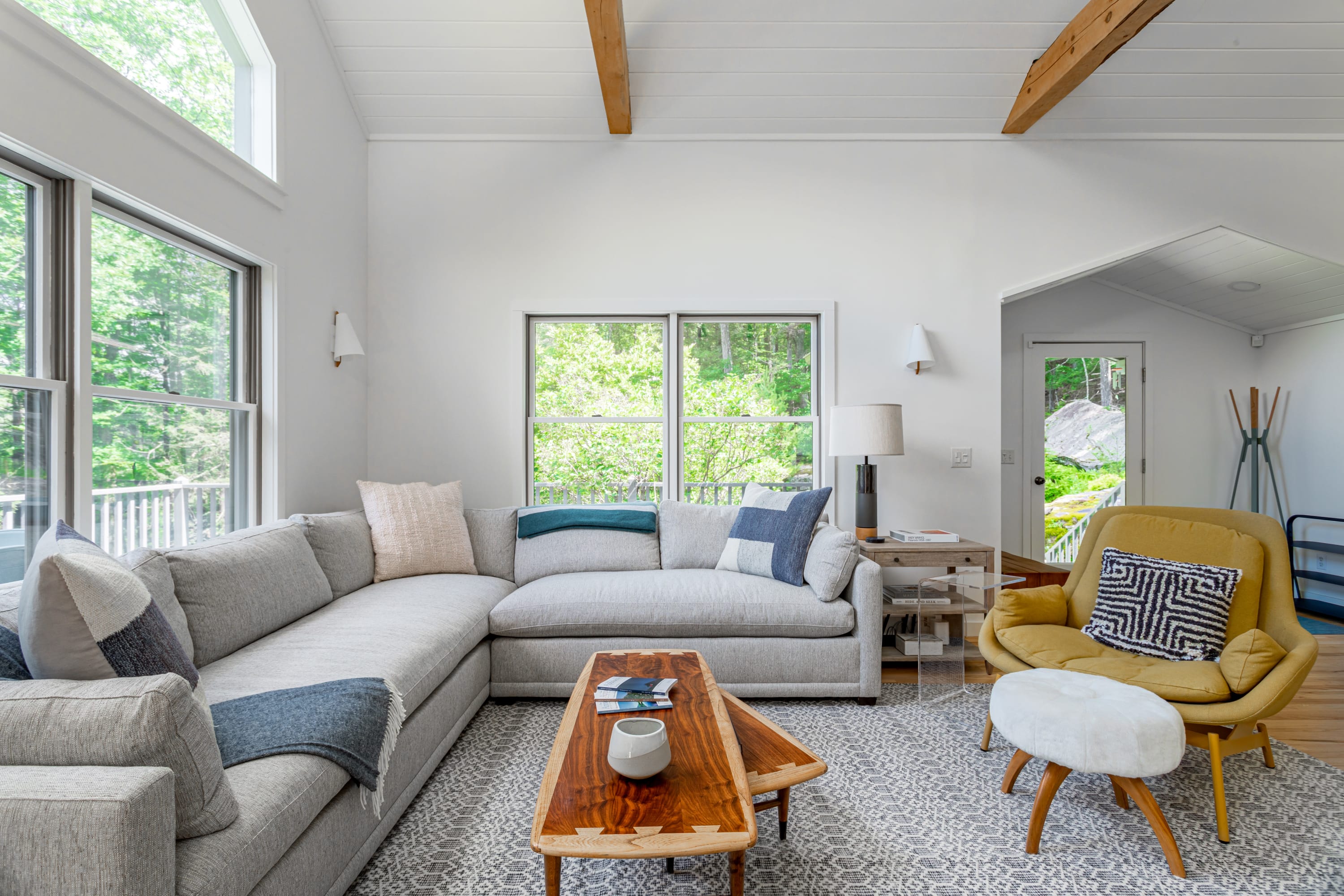 Bright living room with a large gray sectional sofa adorned with throw pillows, a yellow armchair with an ottoman, and a wooden coffee table, all on a patterned area rug.