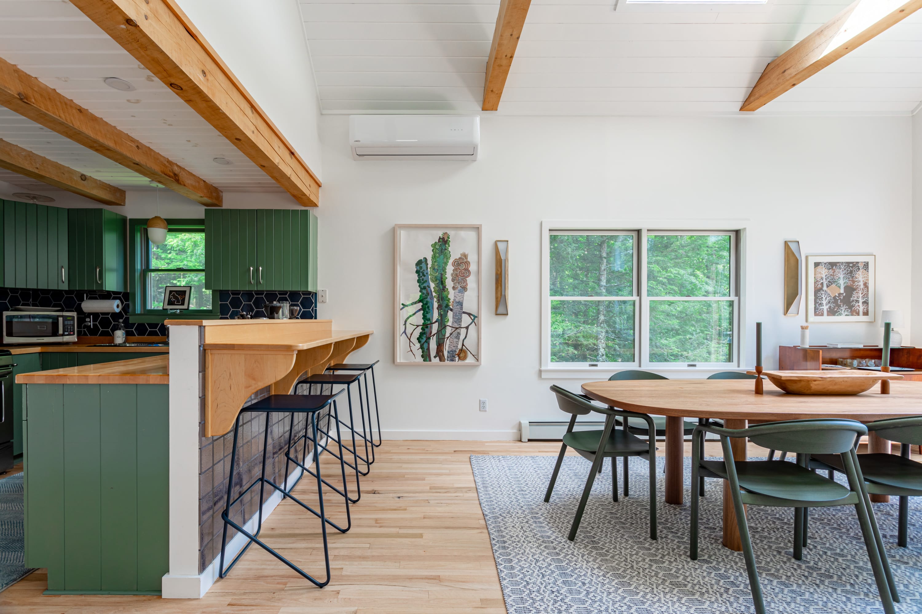 The dining area features a large oval wooden table with dark green chairs, adjacent to a kitchen bar with black stools, all set against white walls and exposed wooden beams, with views of the surrounding greenery.