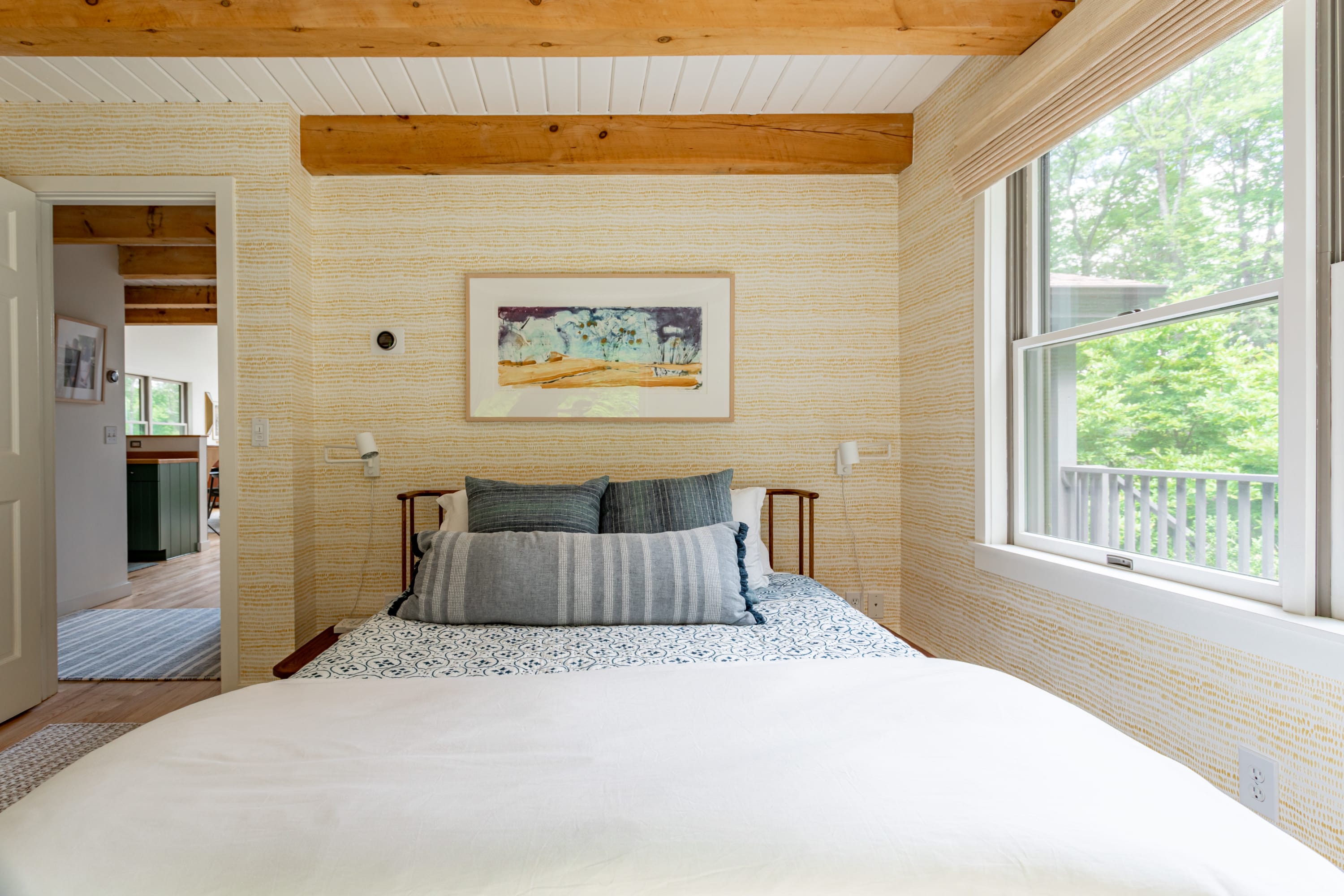 A cozy bedroom featuring yellow patterned wallpaper, exposed wooden ceiling beams, and a bed with blue and grey bedding. A large window offers views of lush greenery outside.