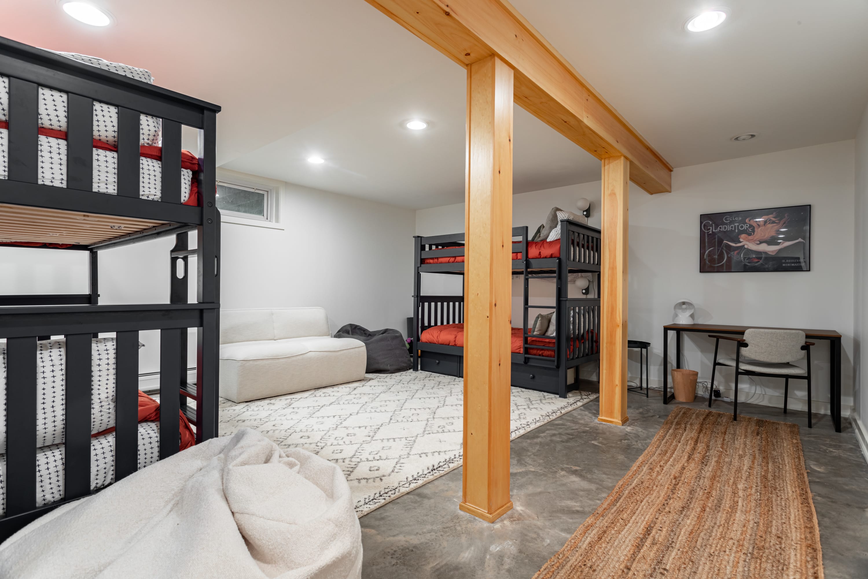 Spacious bunk room featuring two black bunk beds, a white sofa, and a large patterned rug on a concrete floor. Exposed wooden support beams add a rustic touch, and a small desk area is tucked into the corner.