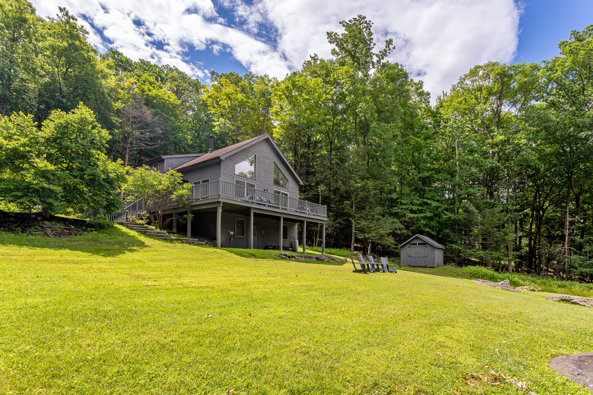 A modern chalet-style house with gray siding and a raised deck, nestled into a lush green hillside surrounded by dense forest. A manicured lawn slopes down from the house, with a small shed and Adirondack chairs visible.