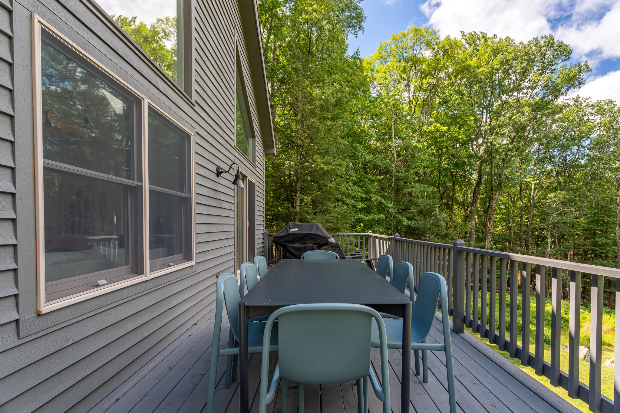 A spacious outdoor deck with gray composite decking, featuring a large black dining table surrounded by light blue chairs. A black grill is visible in the background, and the deck is enclosed by a gray railing, overlooking a lush green forest.