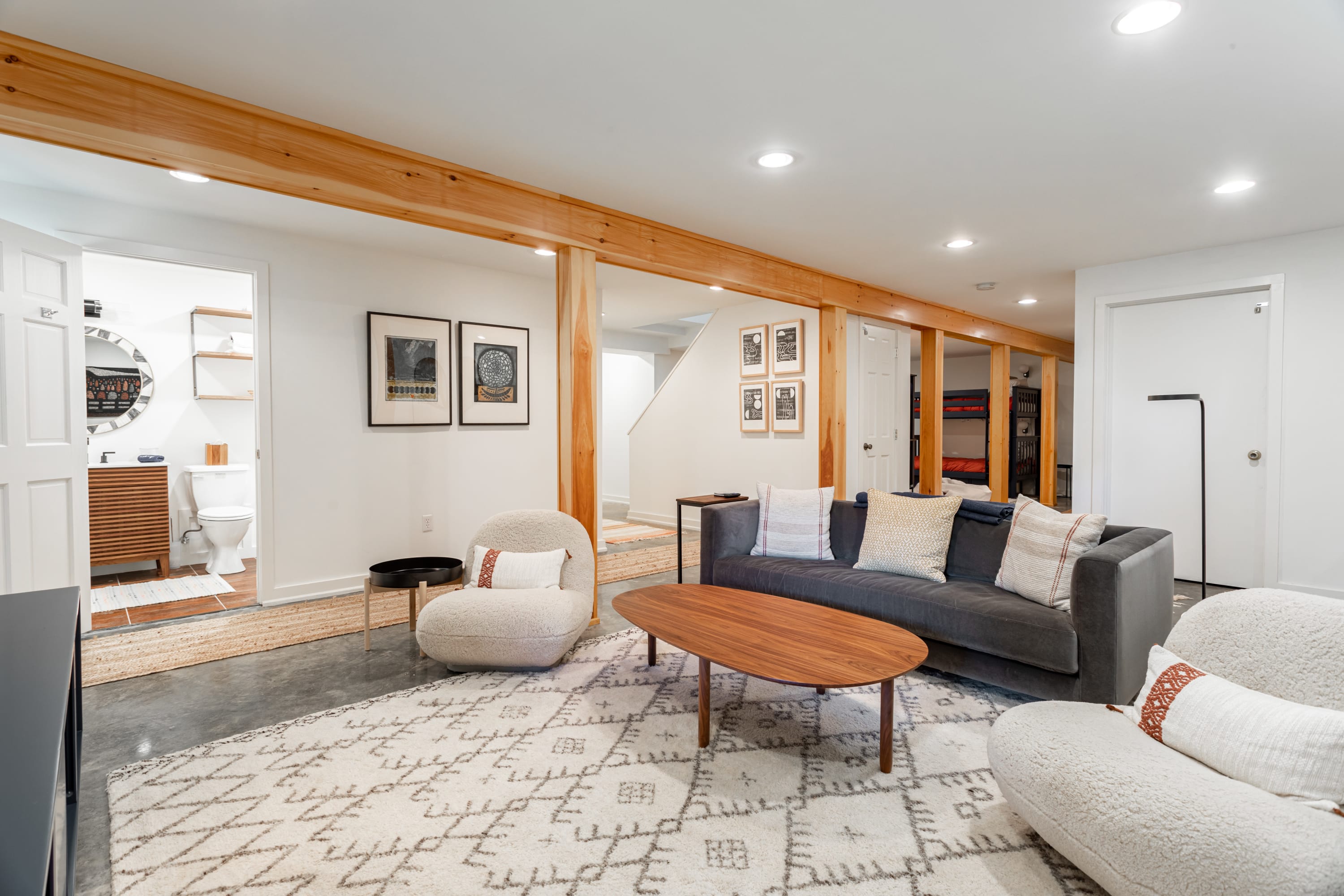 Spacious living room featuring a dark gray sofa, two cream-colored armchairs, and a wooden coffee table on a large patterned rug. Exposed wooden beams define the open-concept space, with a staircase visible in the background.