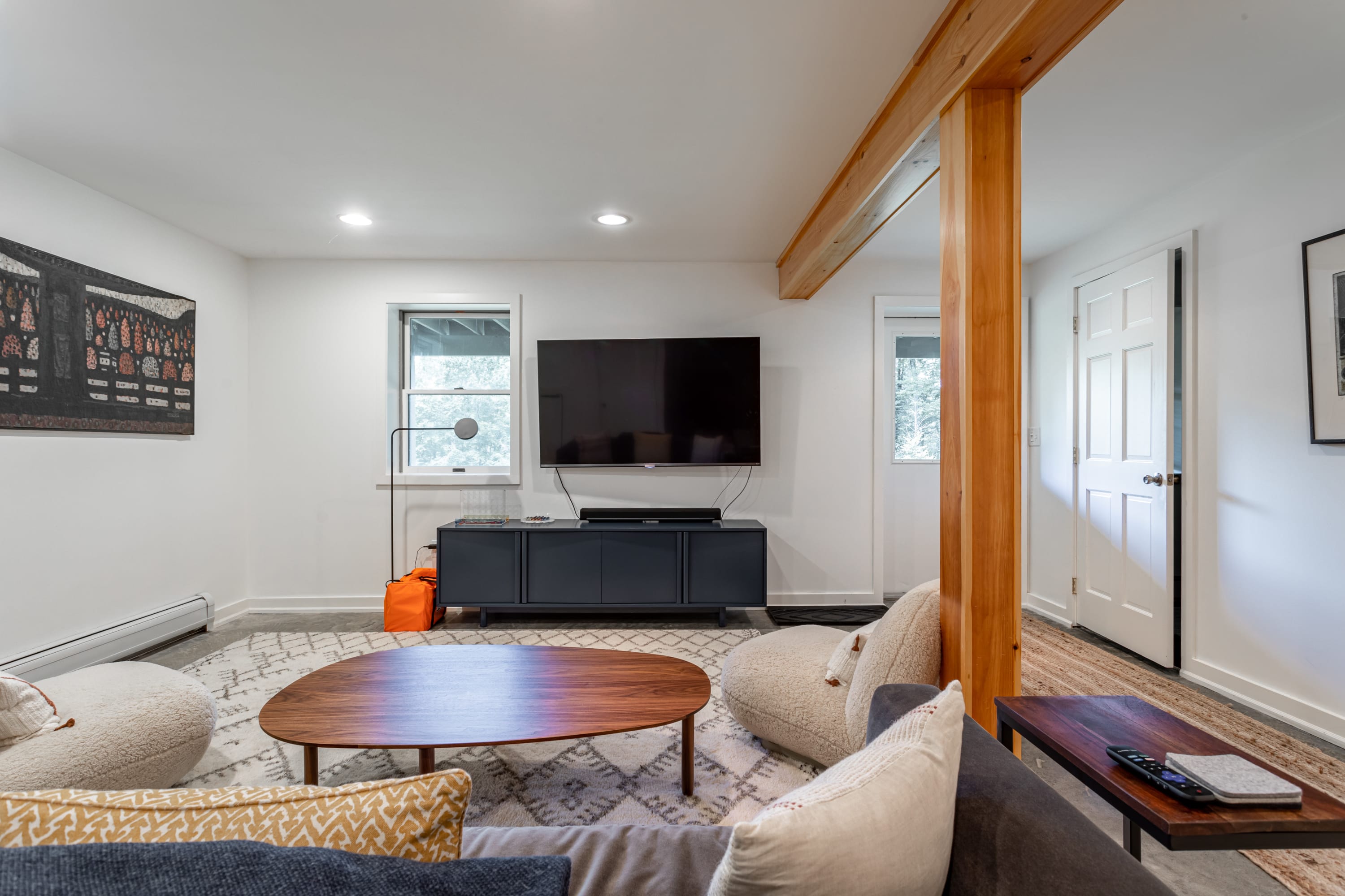 Cozy living room with a large wall-mounted TV above a dark gray media console, a wooden coffee table, and a patterned rug on a concrete floor. A window offers a view of trees, and a prominent wooden beam crosses the ceiling.