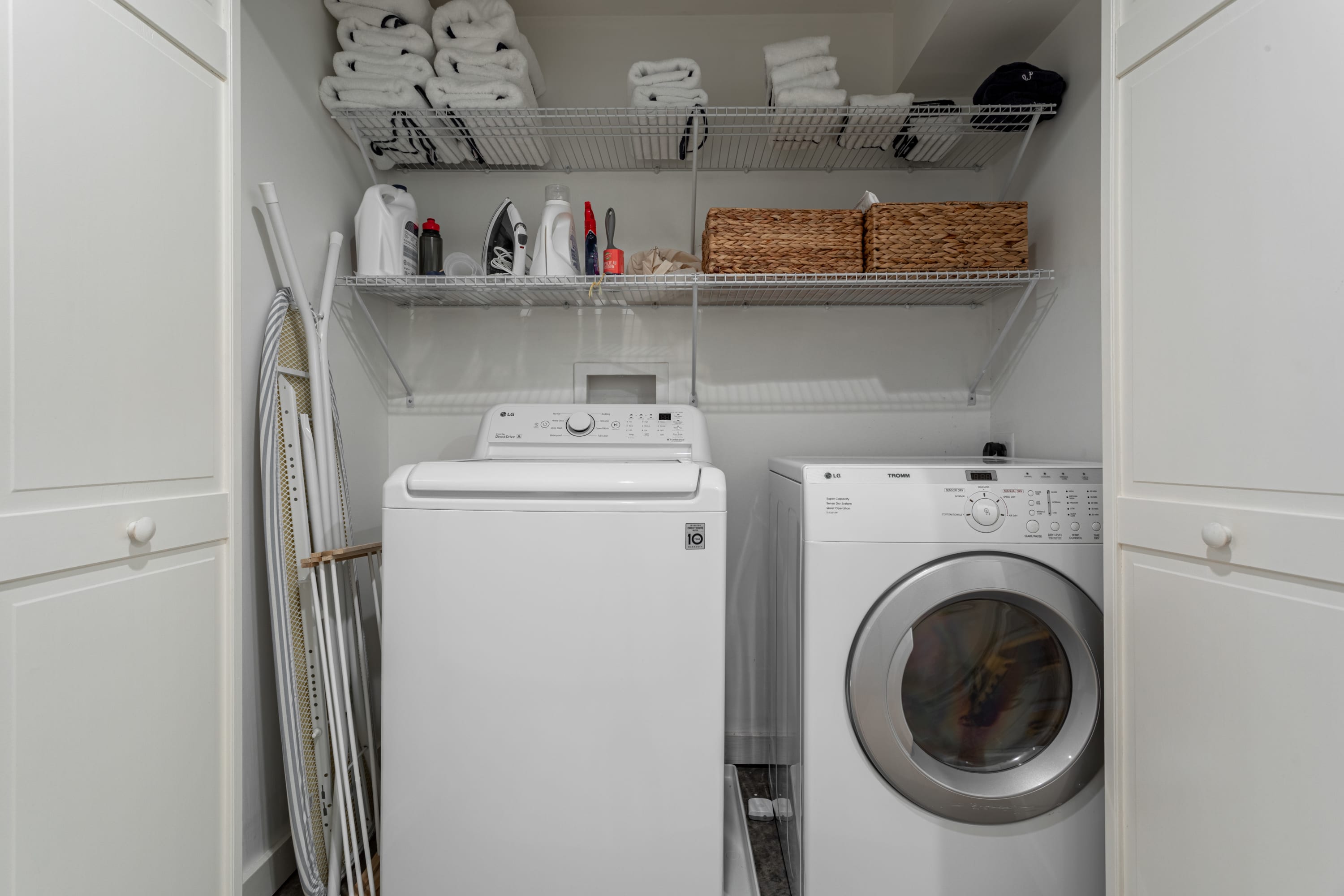 Compact laundry closet featuring a white LG washing machine and a matching LG dryer. Wire shelving above provides storage for neatly stacked towels and cleaning supplies, with an ironing board stored beside the machines.