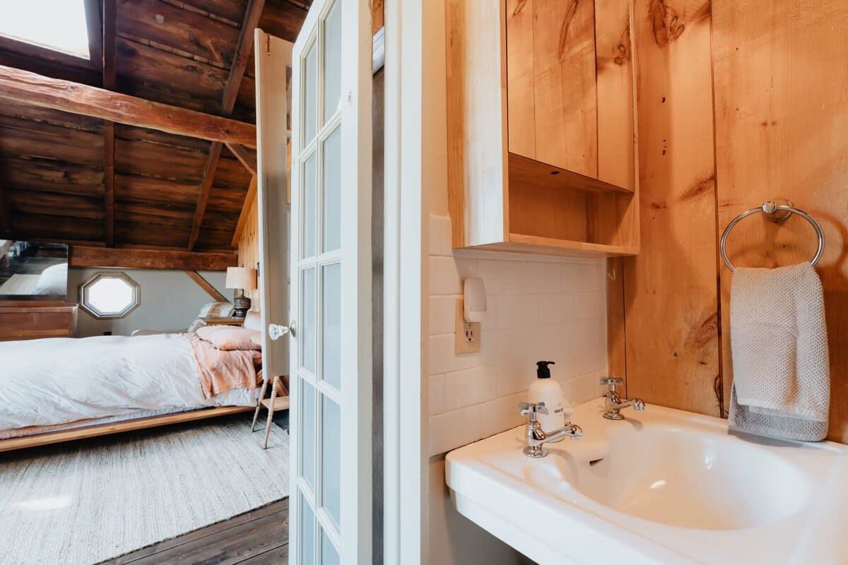 View into a compact bathroom with a white sink and wood-paneled walls, showing a partial view of the adjacent bedroom through a set of white French doors. Exposed wooden beams are visible in the ceiling.