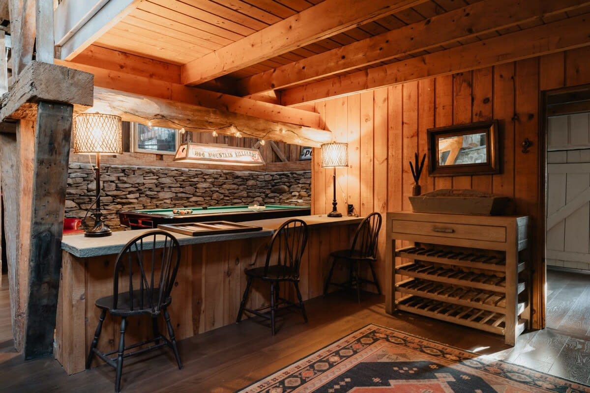 A rustic game room featuring a wooden bar with stools, two table lamps, and a pool table visible in the background against a stone wall. The room has exposed wooden beams and paneling.