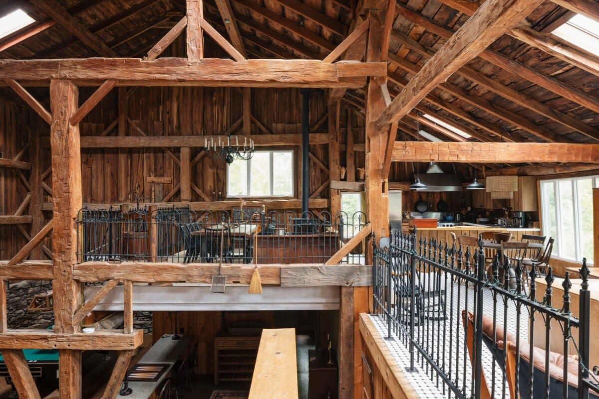 Expansive multi-level interior showcasing the barn's original timber frame construction with exposed wooden beams and plank walls. A dining area and kitchen are visible on the upper level, overlooking a lower floor with a game table.