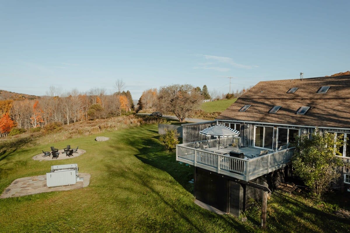 Aerial view of the barn-style house with a large deck overlooking a vast green lawn and distant autumn-colored hills. The property is surrounded by trees and features an outdoor seating area and fire pit.