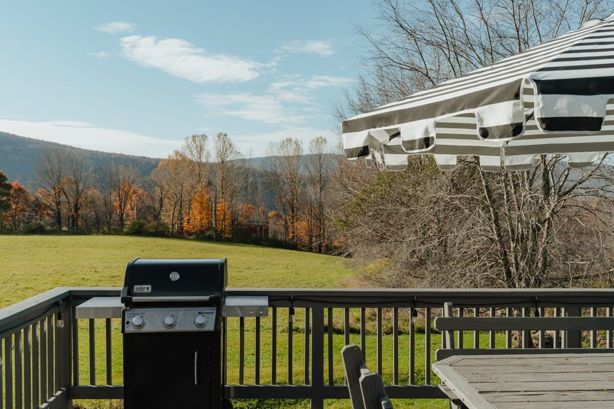 A close-up view of the outdoor deck with a black BBQ grill and a striped umbrella, showcasing a vibrant autumn landscape with colorful trees and rolling hills in the background under a clear sky.
