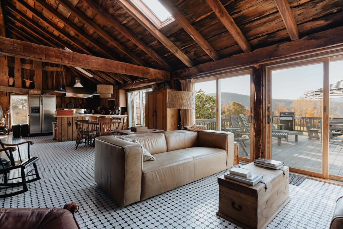 A bright, open-concept living space features a large tan leather sofa, a wooden coffee table, and black and white tiled floors. Large sliding glass doors open to a deck, offering expansive views of autumn-colored mountains.