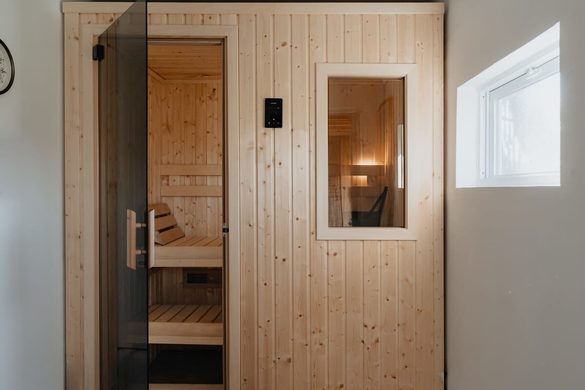 A modern wooden sauna with a glass door and a small window, featuring light wood paneling and tiered benches inside. A control panel is mounted on the wall.