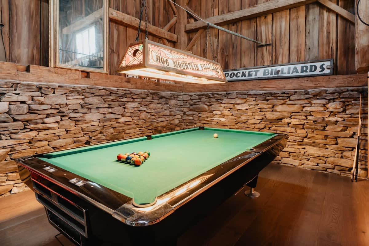 A rustic game room boasts a green-felt pool table under a vintage "Hog Mountain Billiards" light fixture, surrounded by stone walls and exposed wooden beams, with cues leaning against the wall.