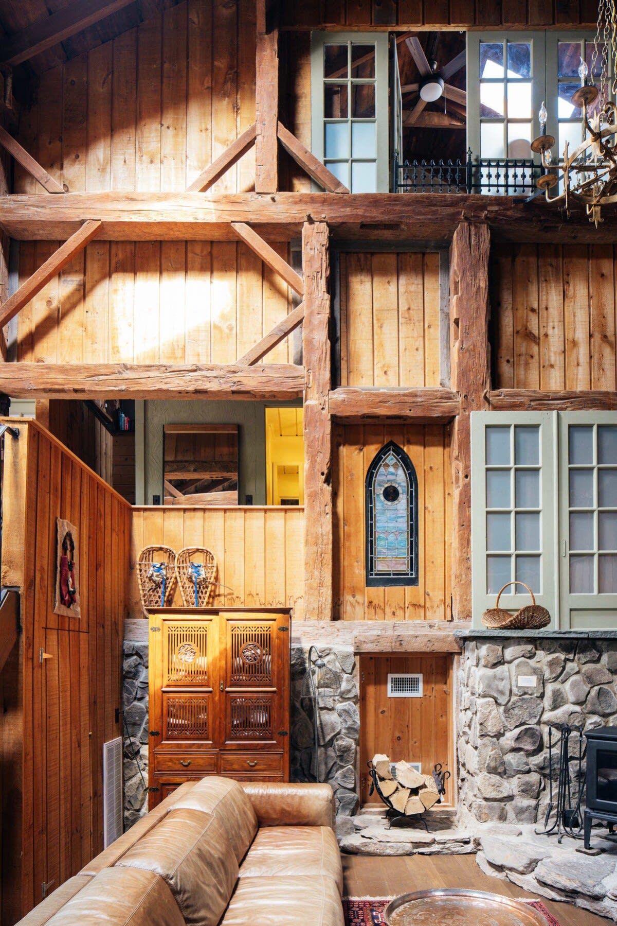 Grand living space showcasing a multi-level design with extensive wood paneling and exposed timber beams. A stone wall with a wood-burning stove is visible, along with a leather sectional sofa and unique architectural details like a stained-glass window.