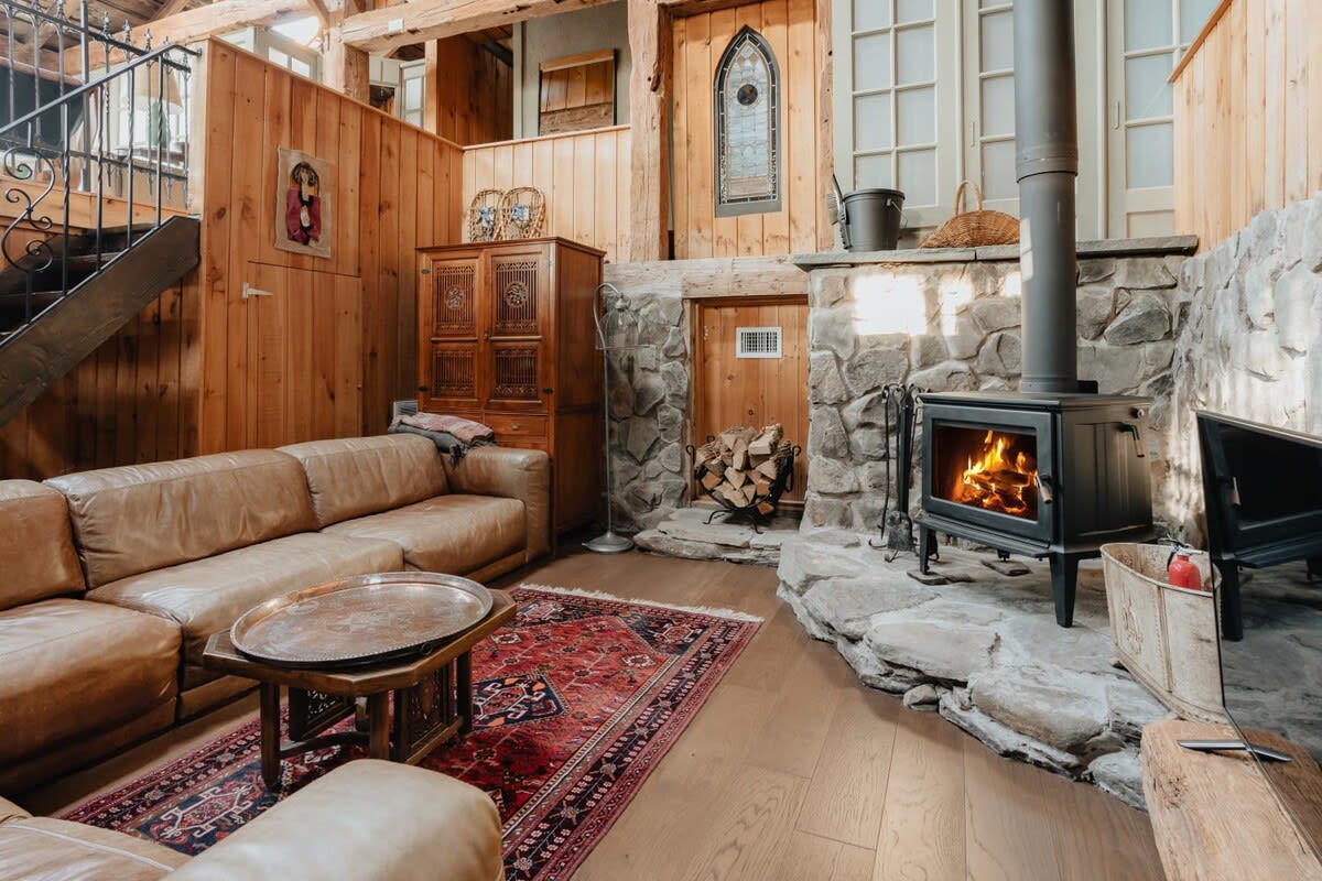 Spacious living room with a large leather sectional sofa, a rustic wood-burning stove set against a stone hearth, and warm wood-paneled walls. A red patterned rug adds a pop of color to the hardwood floor.