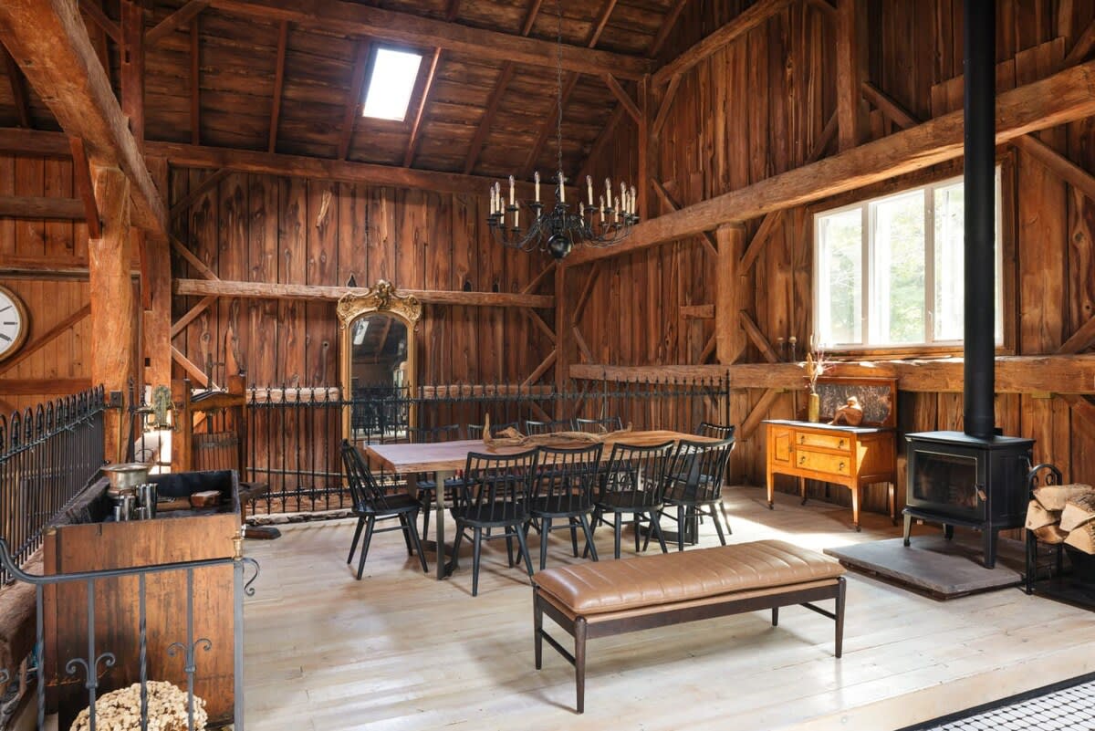Expansive dining room with a long wooden table and numerous black chairs, illuminated by a large black chandelier. The room features exposed wooden beams, a wood-burning stove, and a large ornate mirror.
