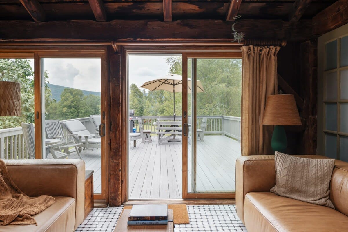 Living room with two light brown sofas facing large sliding glass doors that open onto a spacious deck with outdoor furniture and distant mountain views. The room features a wooden ceiling and tiled floor.